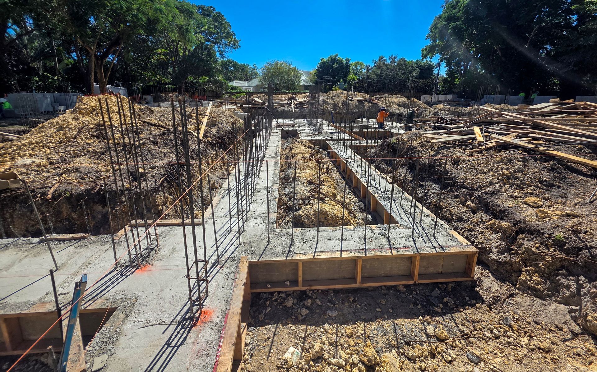 Construction site: concrete foundation forms with rebar, dirt, and a worker under a blue sky.