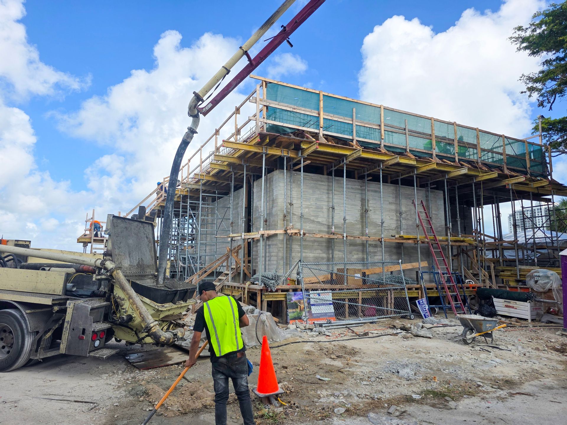 Concrete construction site; a worker in a safety vest sweeps debris. A pump truck pours concrete into the building's framework.
