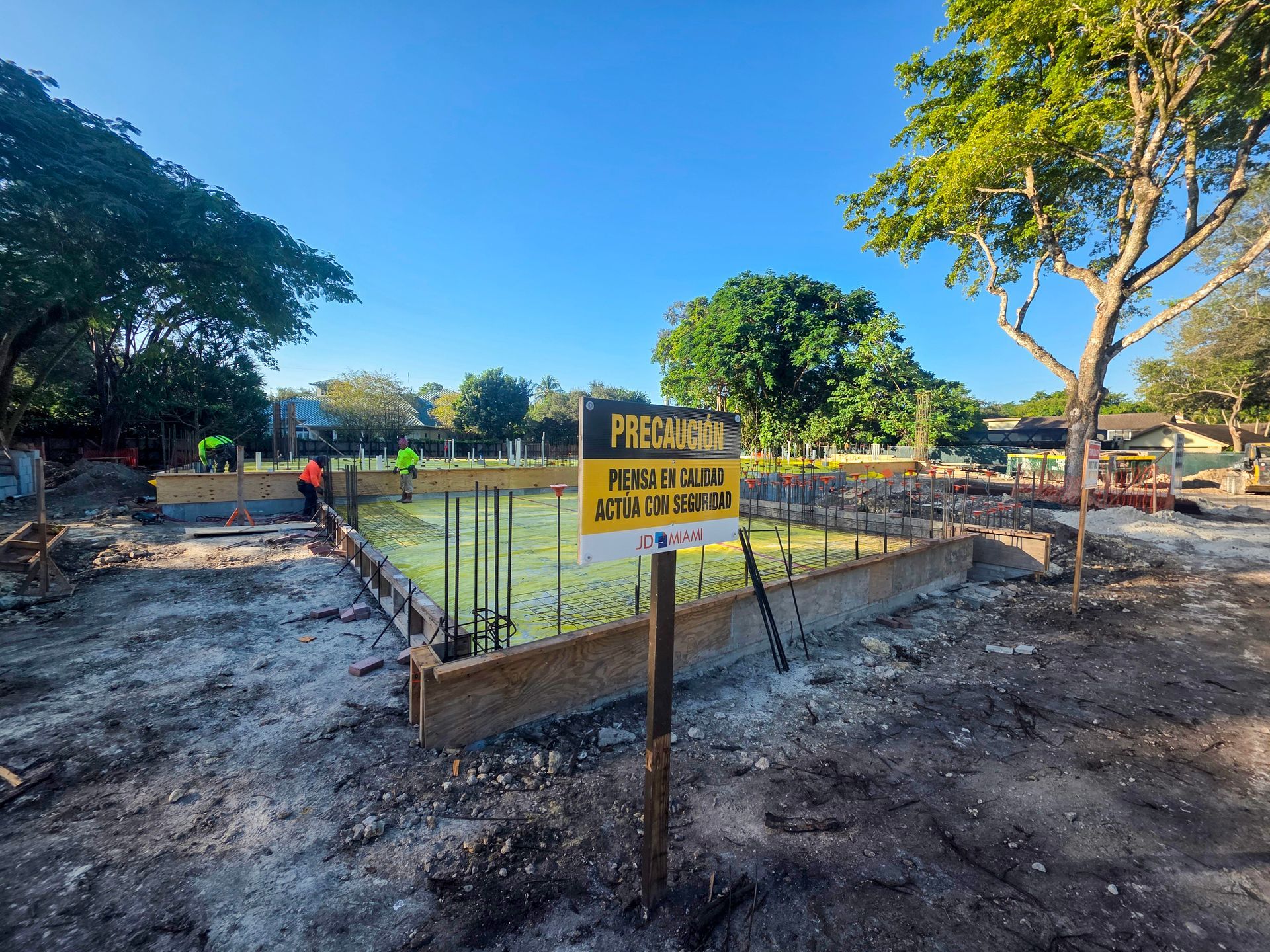 Construction site with foundation, rebar, and sign under a blue sky. Workers visible in the background.