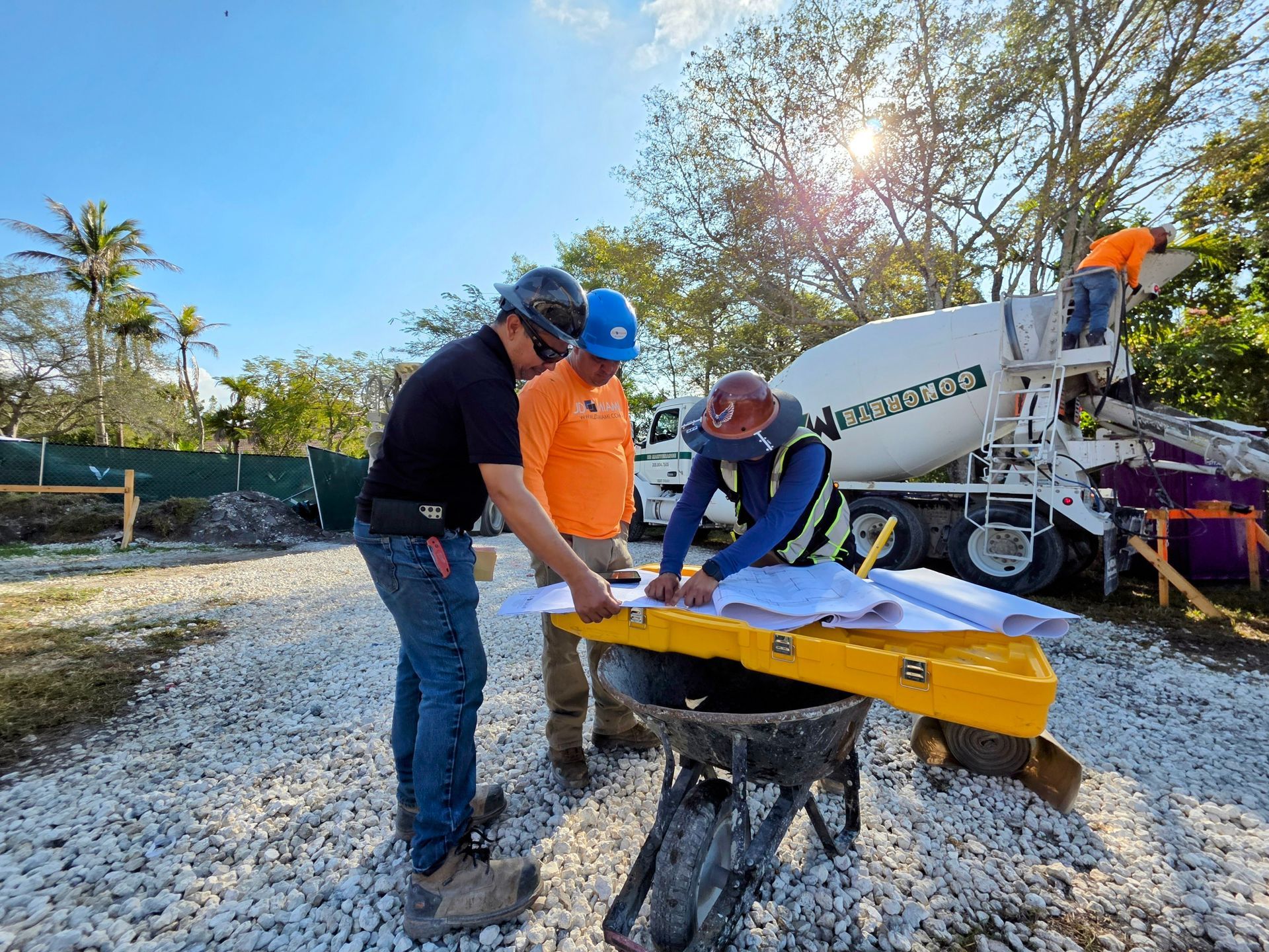 Construction workers review blueprints outdoors near a cement truck; bright sun.