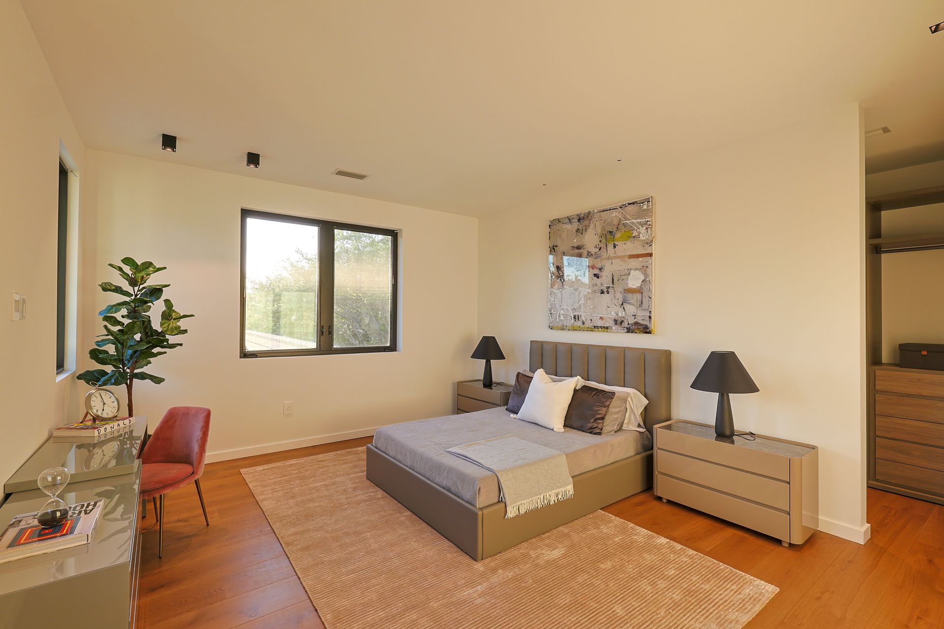 Bedroom with bed, nightstands, desk, chair, and large window. Neutral color scheme, wood flooring.