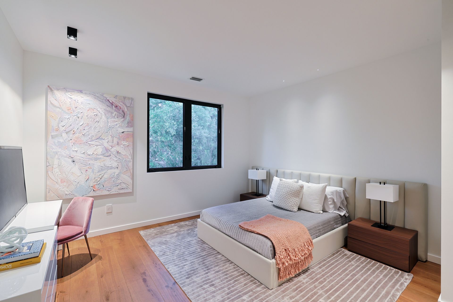 Bedroom with bed, desk, art, and window. White walls, wooden floor, neutral tones.