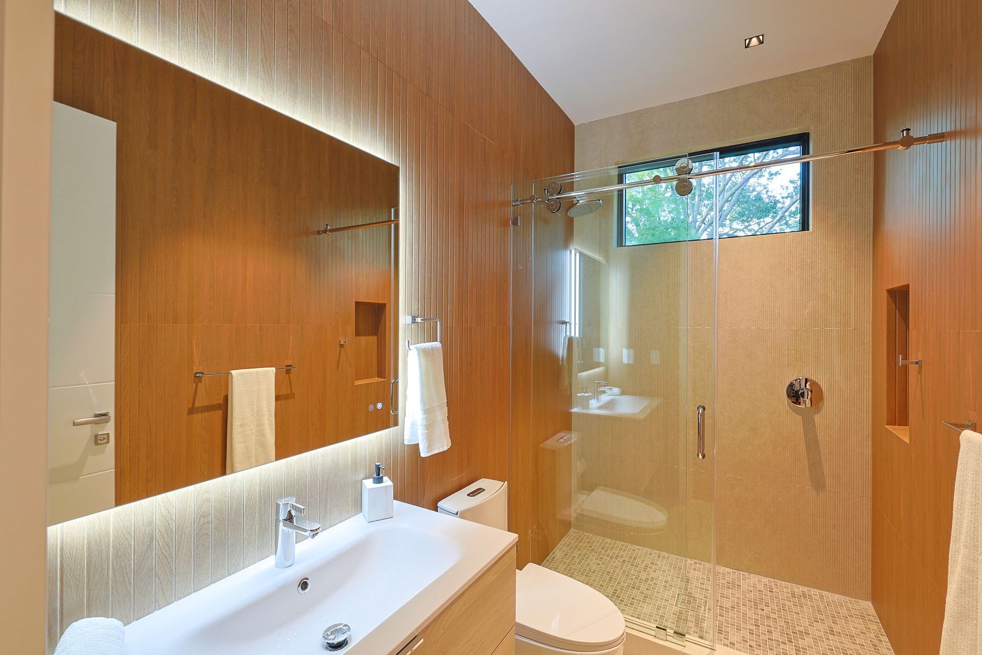 Modern bathroom with wood paneling, large mirror, glass shower, and pebble floor.