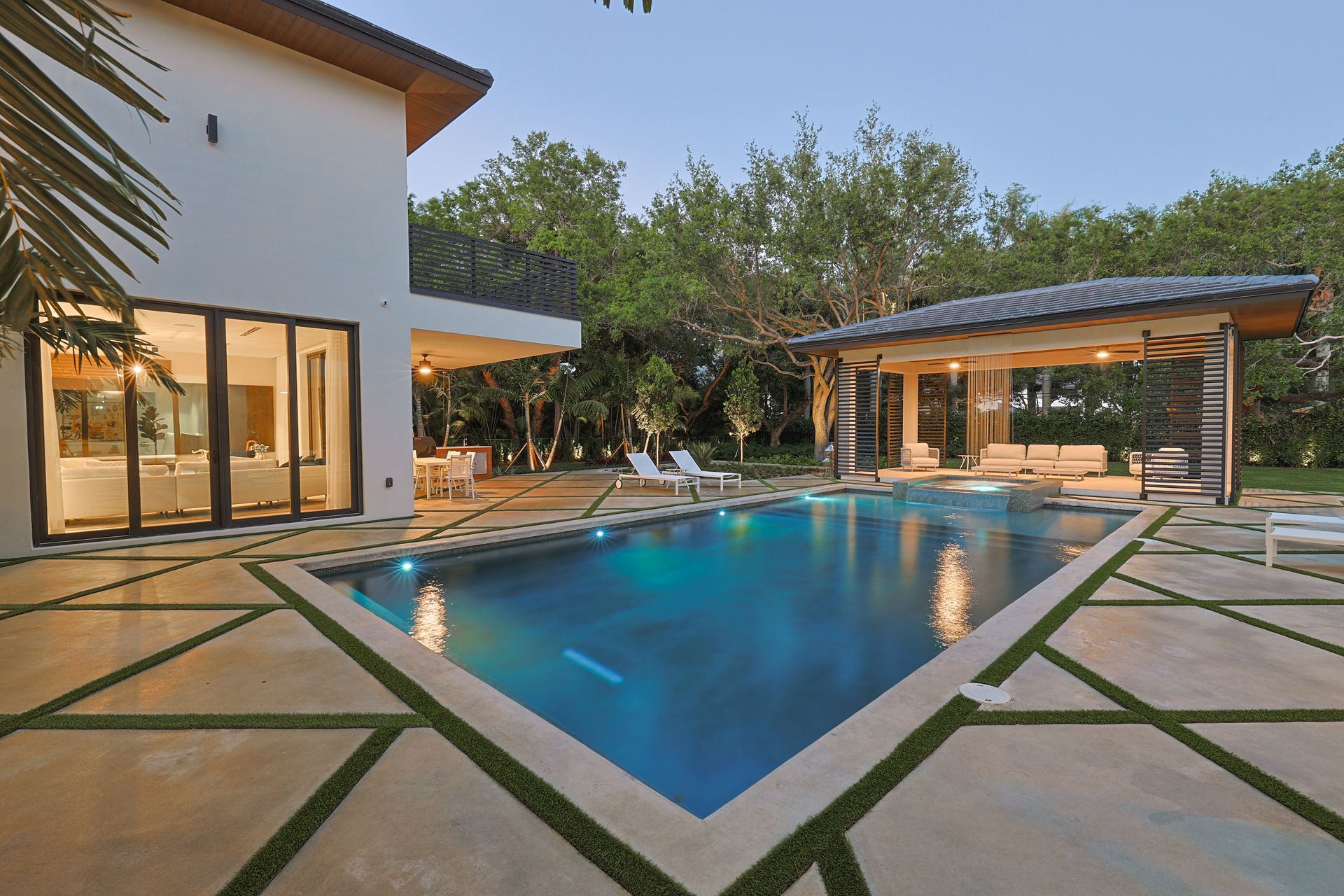 Swimming pool at twilight with a gazebo, lit house, and concrete patio.