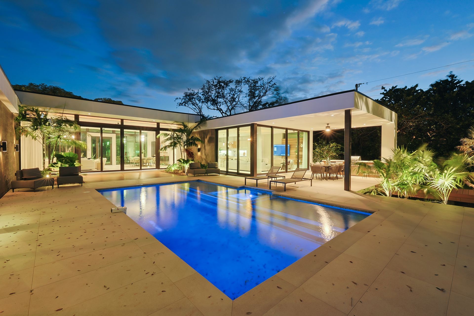 Modern home with a blue swimming pool, illuminated at dusk. Features include a patio, outdoor dining, and folding glass doors.