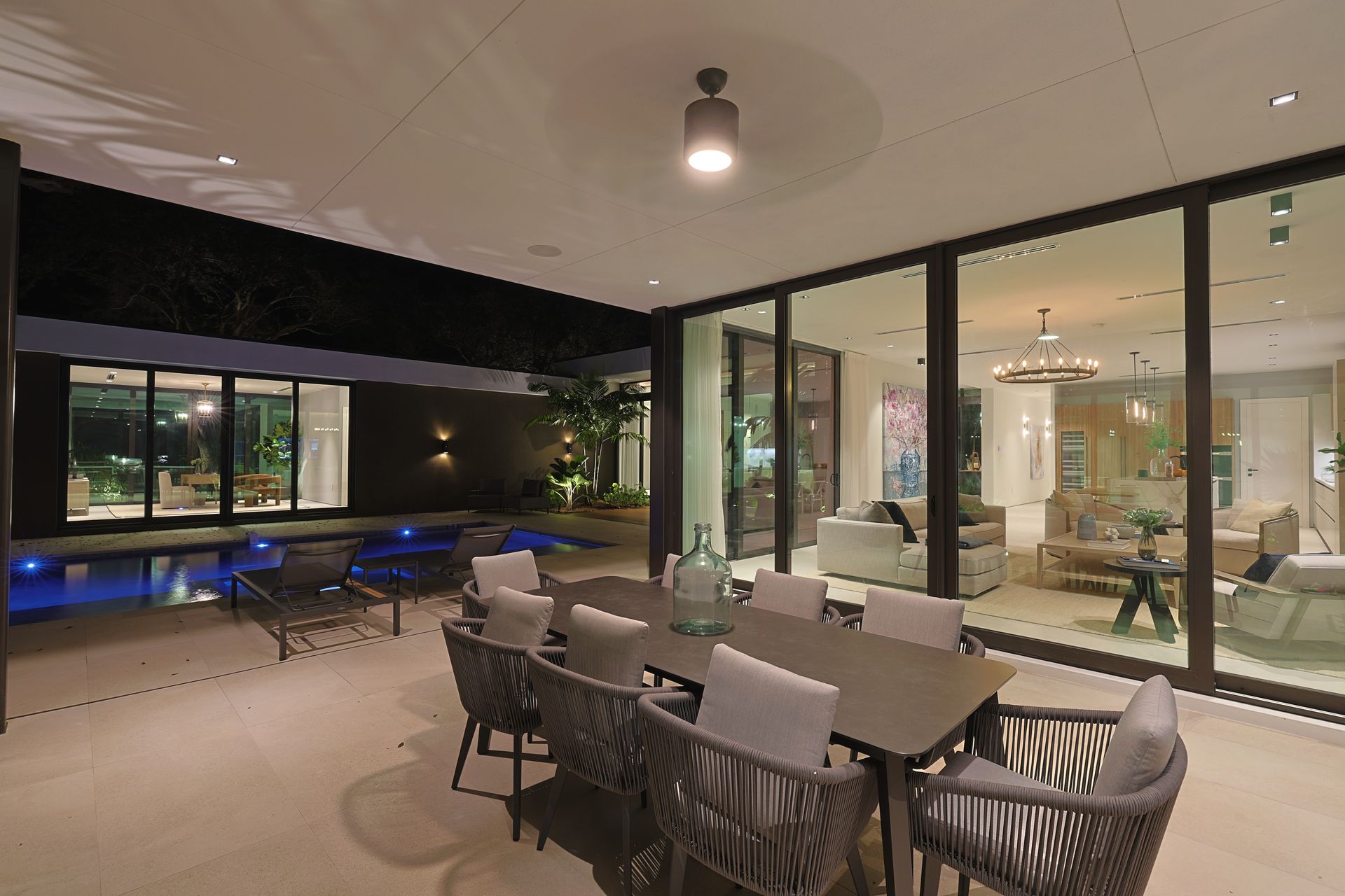 Outdoor dining area with table and chairs next to glass wall, pool visible in background at night.