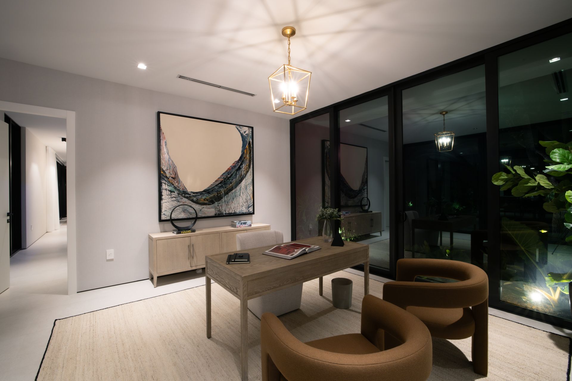 A modern home office with a desk, chairs, artwork, and large windows looking out at night.