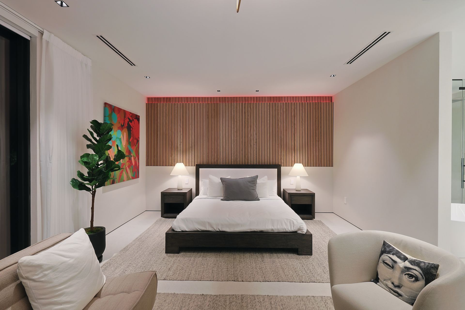 Bedroom with bed, art, plants, and seating. Neutral tones with accent lighting.