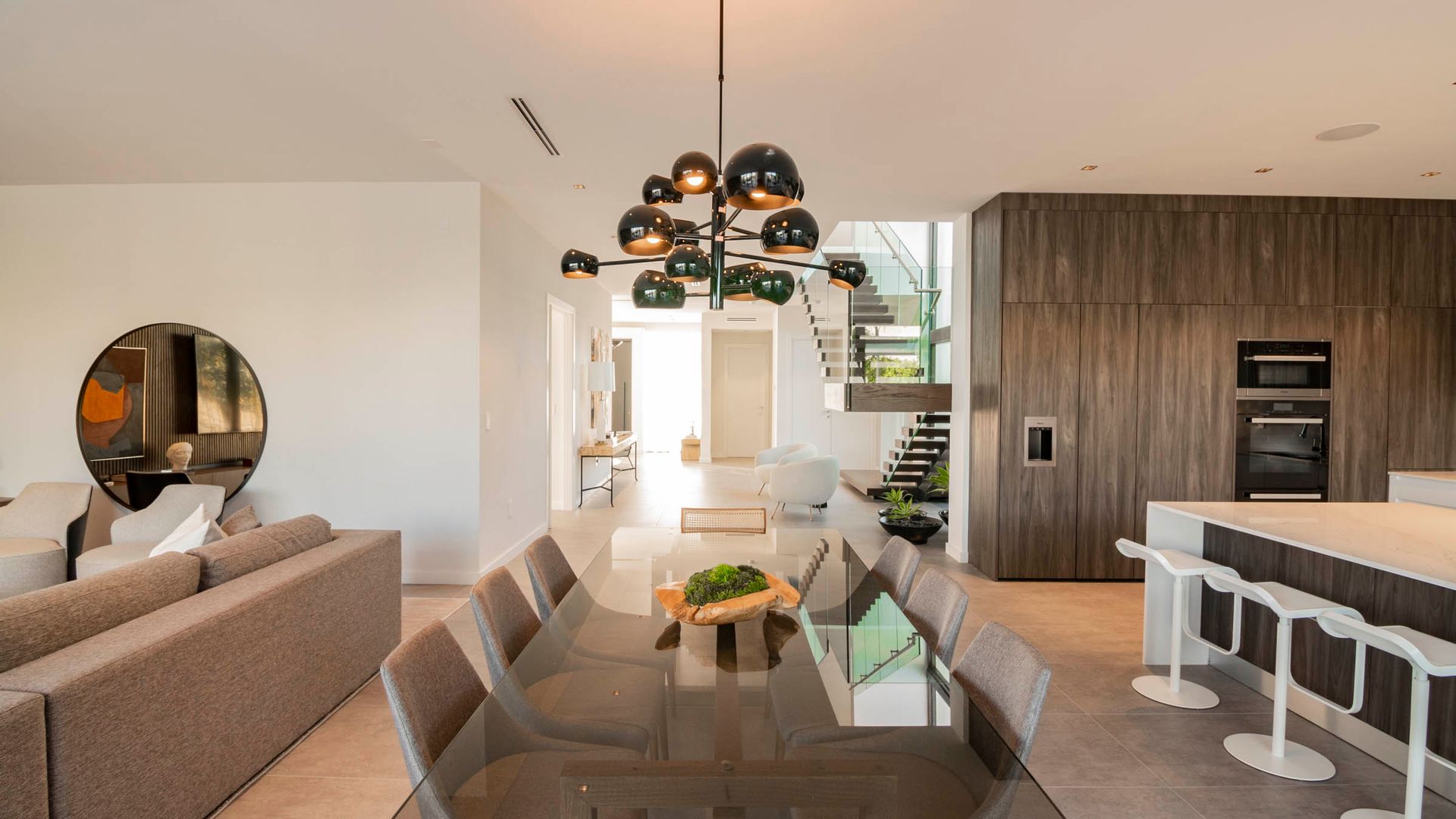 Modern dining room with a long table, chandelier, and adjacent kitchen with bar seating.