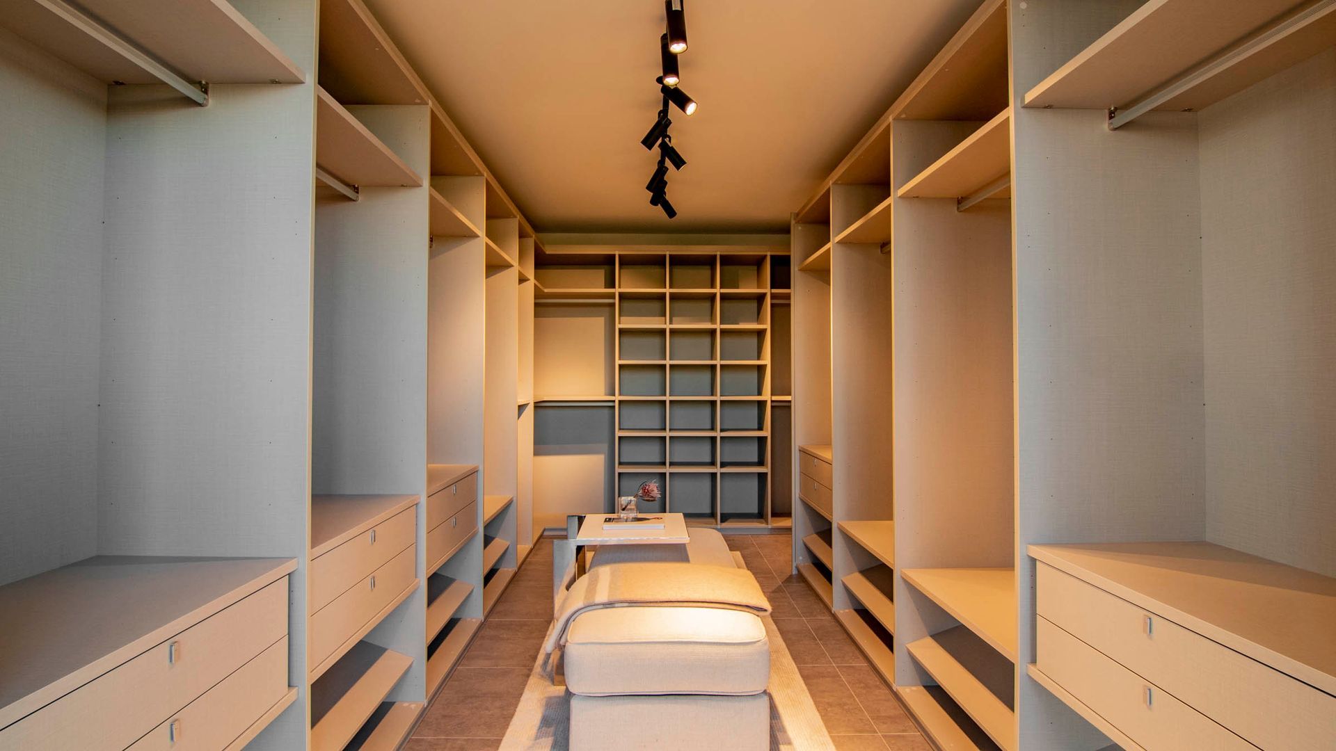 Empty walk-in closet with beige shelves, drawers, and a central bench; lit by track lighting.
