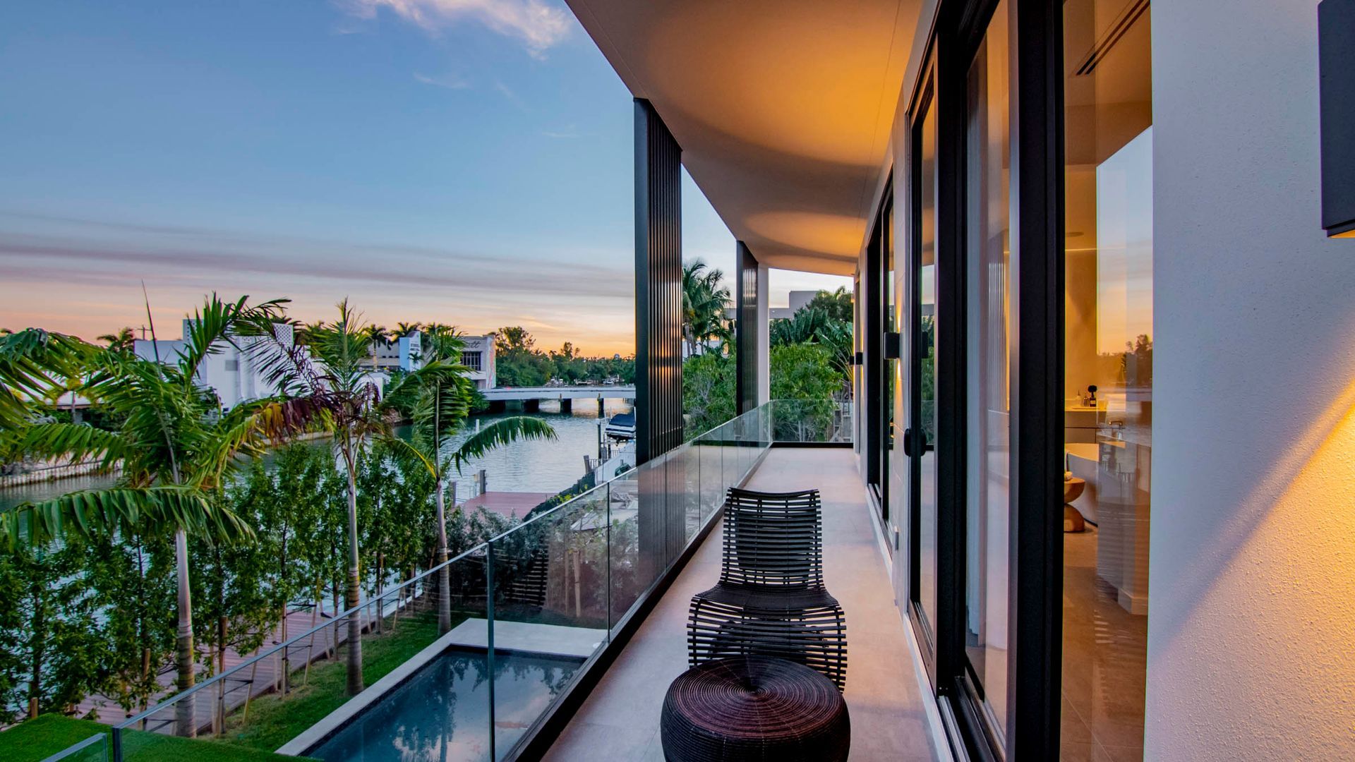 Balcony overlooking water, featuring seating, glass walls, and trees under a colorful sky.