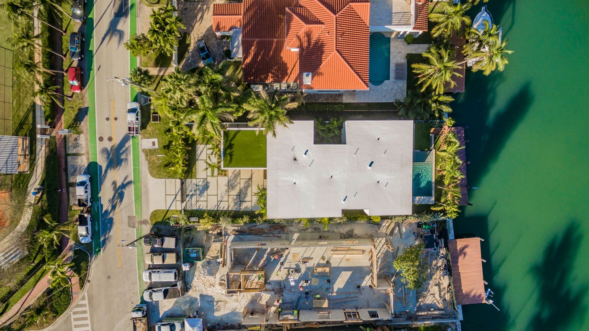 Aerial view of waterfront houses with docks, pools, and lush green landscaping. A construction site is at the bottom.