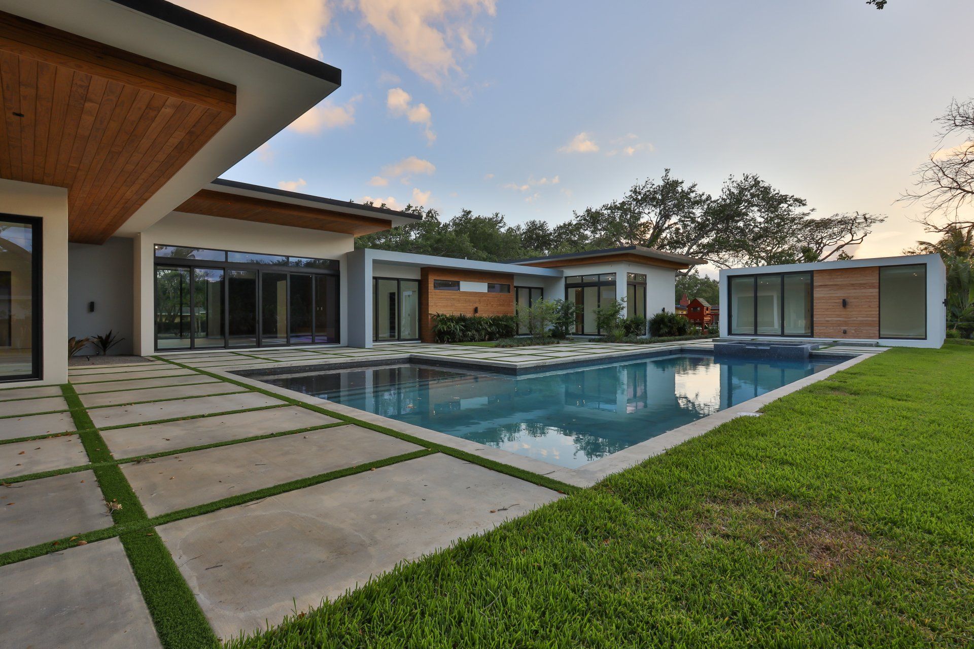 Modern home with a pool, concrete patio, and grass lawn. Clear blue water reflects the sky.