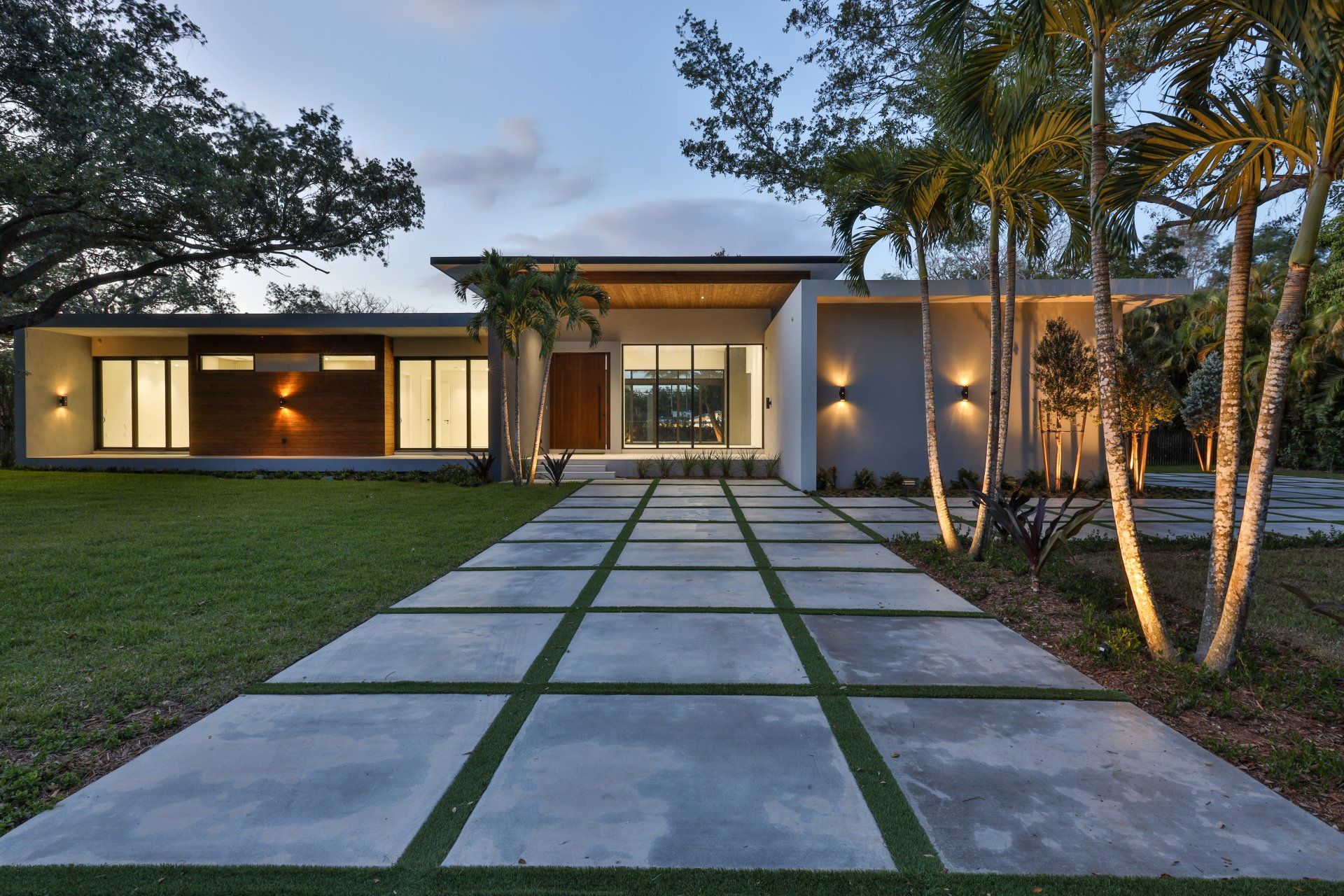 Modern home with concrete walkway, green grass, and palm trees at dusk.