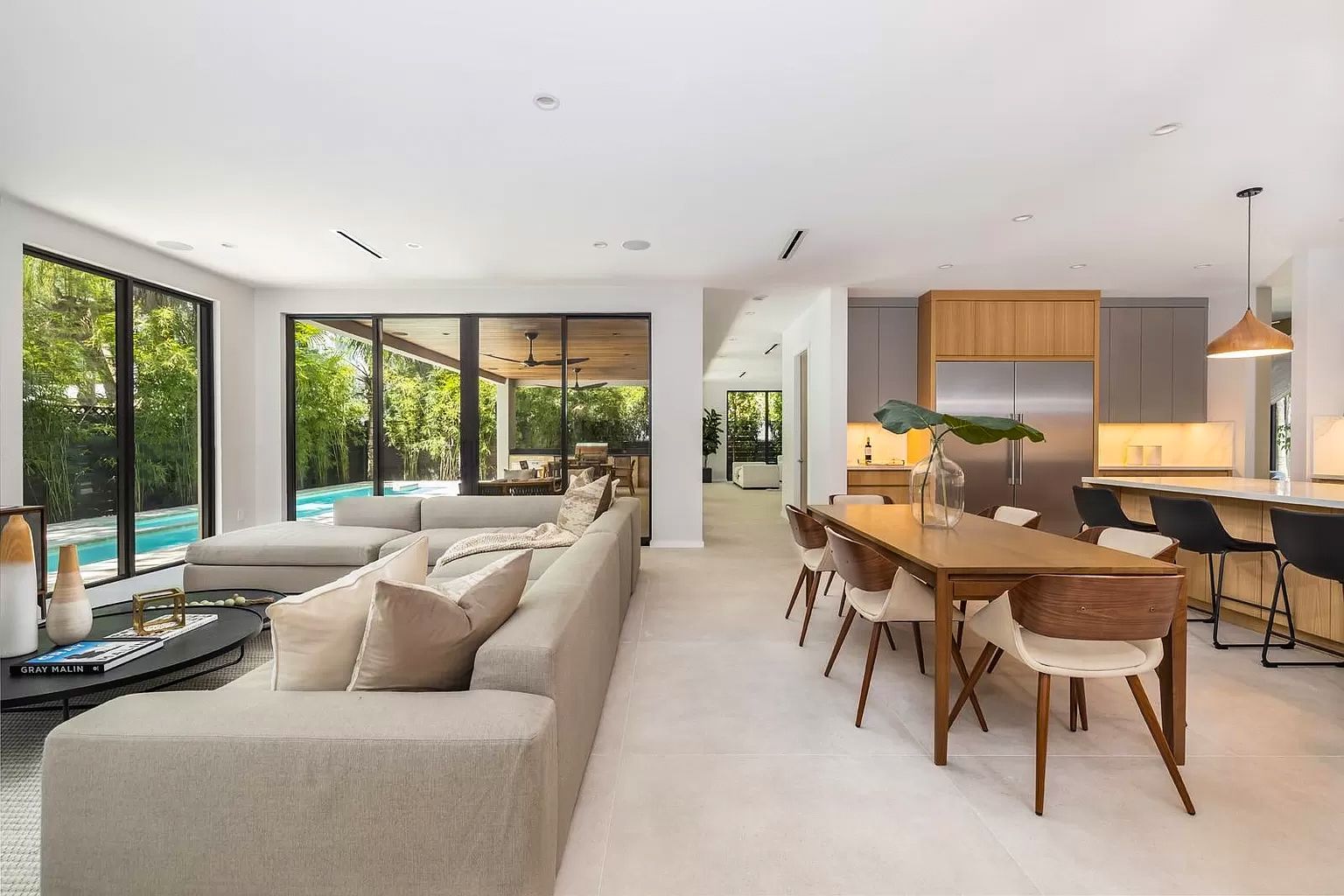 Modern open-plan living space with a large sofa, dining table, and kitchen. Large windows overlook a pool.