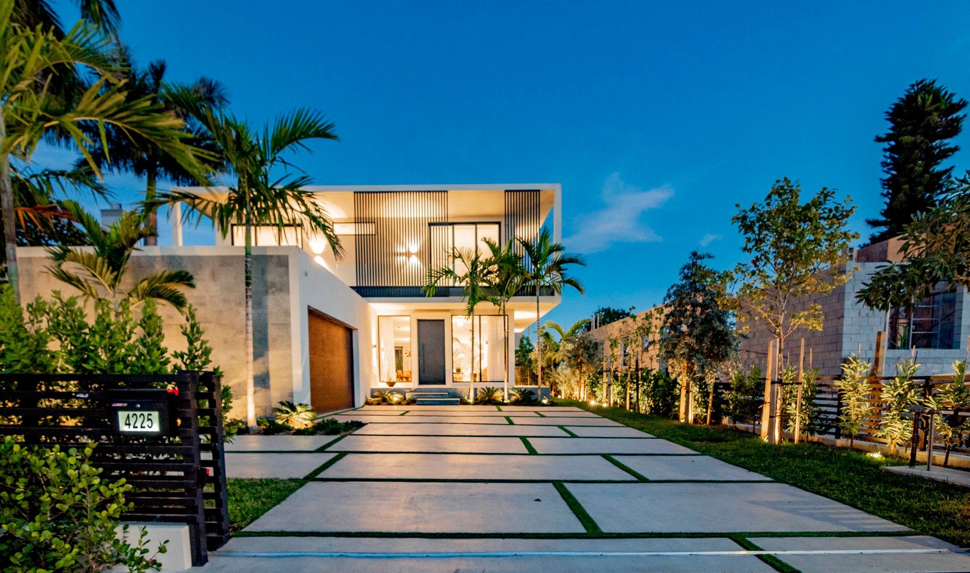 Modern two-story house with a long, lighted driveway, palm trees, and evening sky.