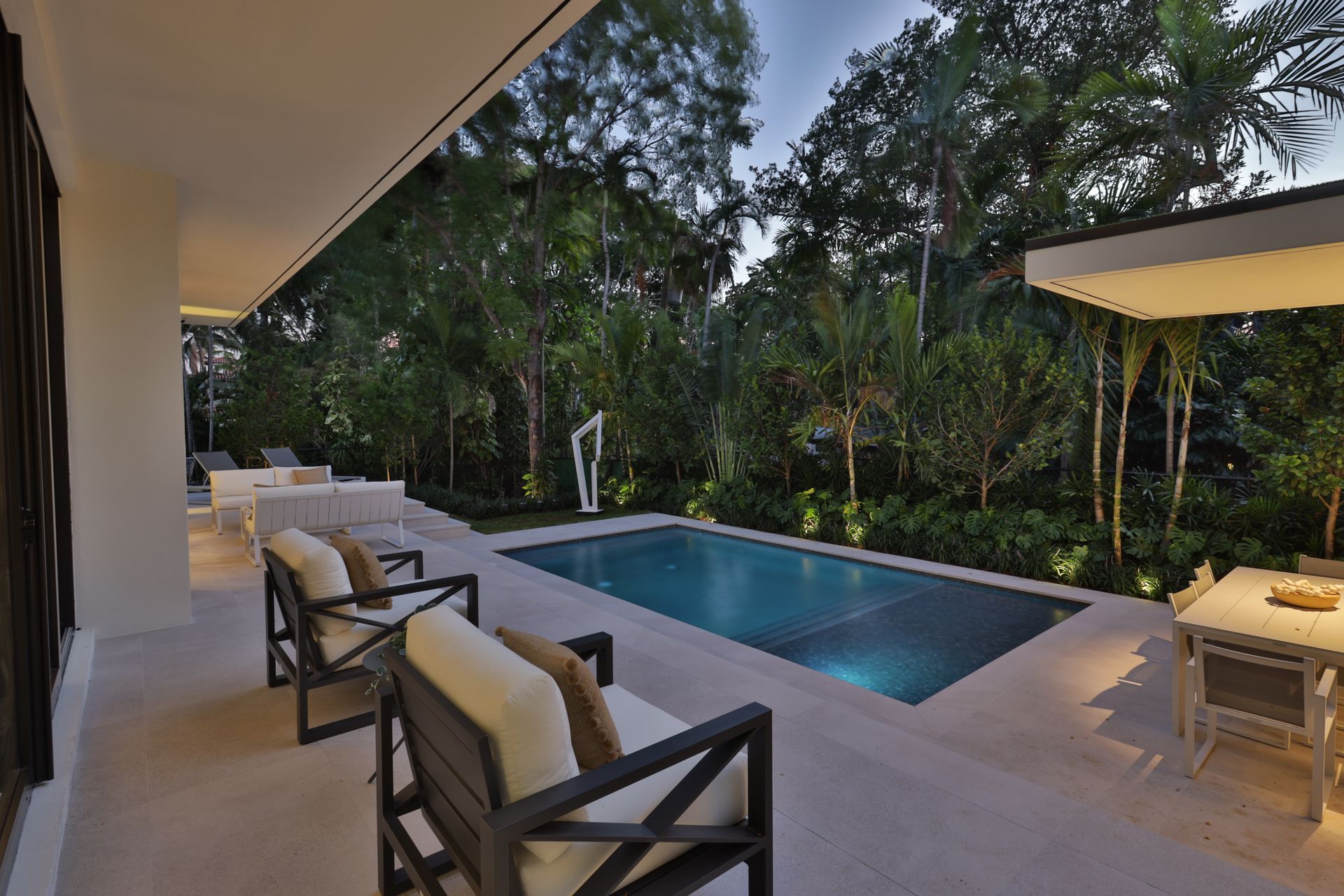 Patio with pool, dark wooden chairs with white cushions, outdoor dining, lush greenery.