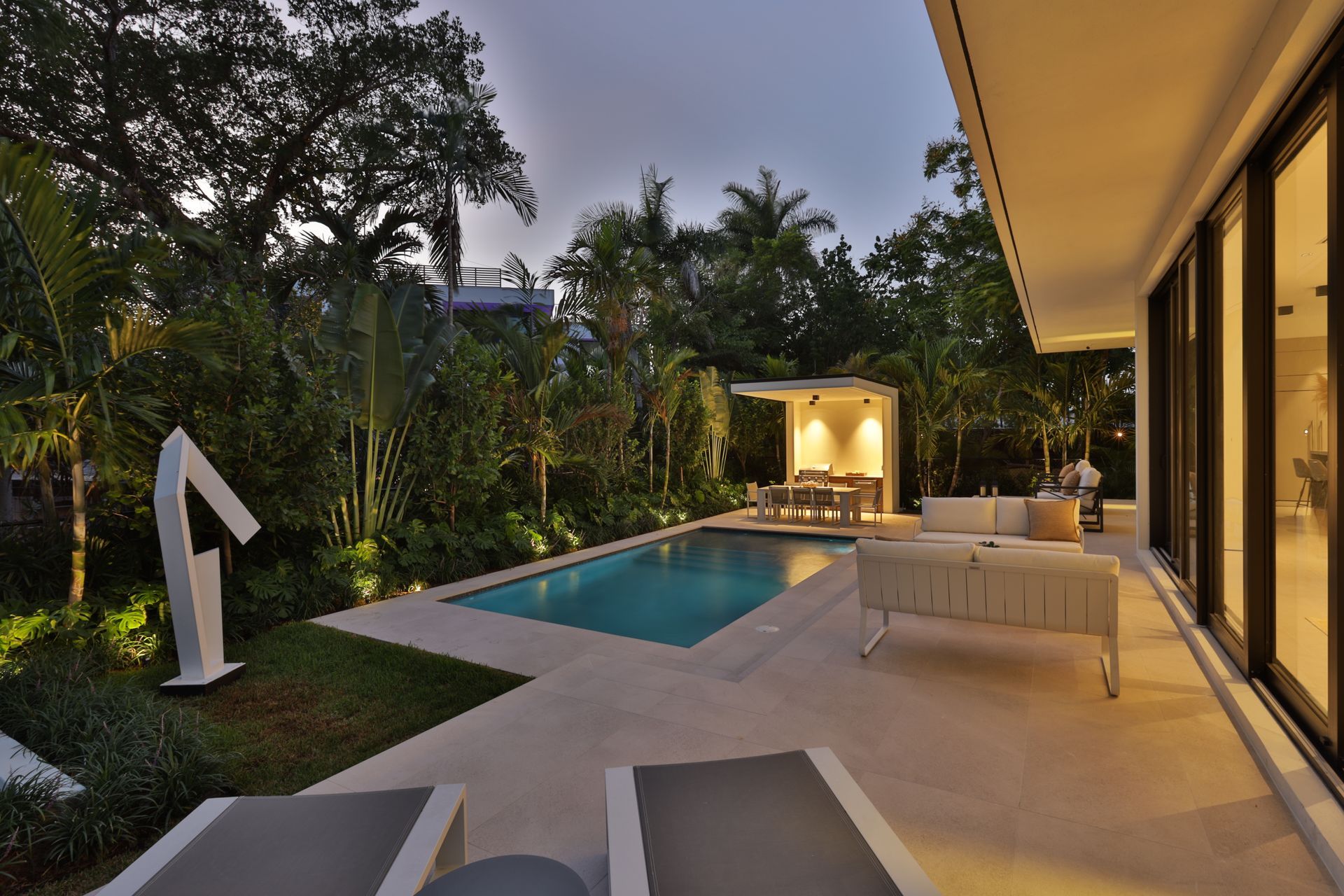 Backyard with pool, patio, and modern house, surrounded by lush greenery.