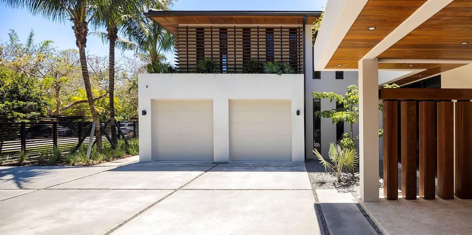 Modern two-story house with a white exterior, two-car garage, and wooden accents; palm trees and driveway in view.