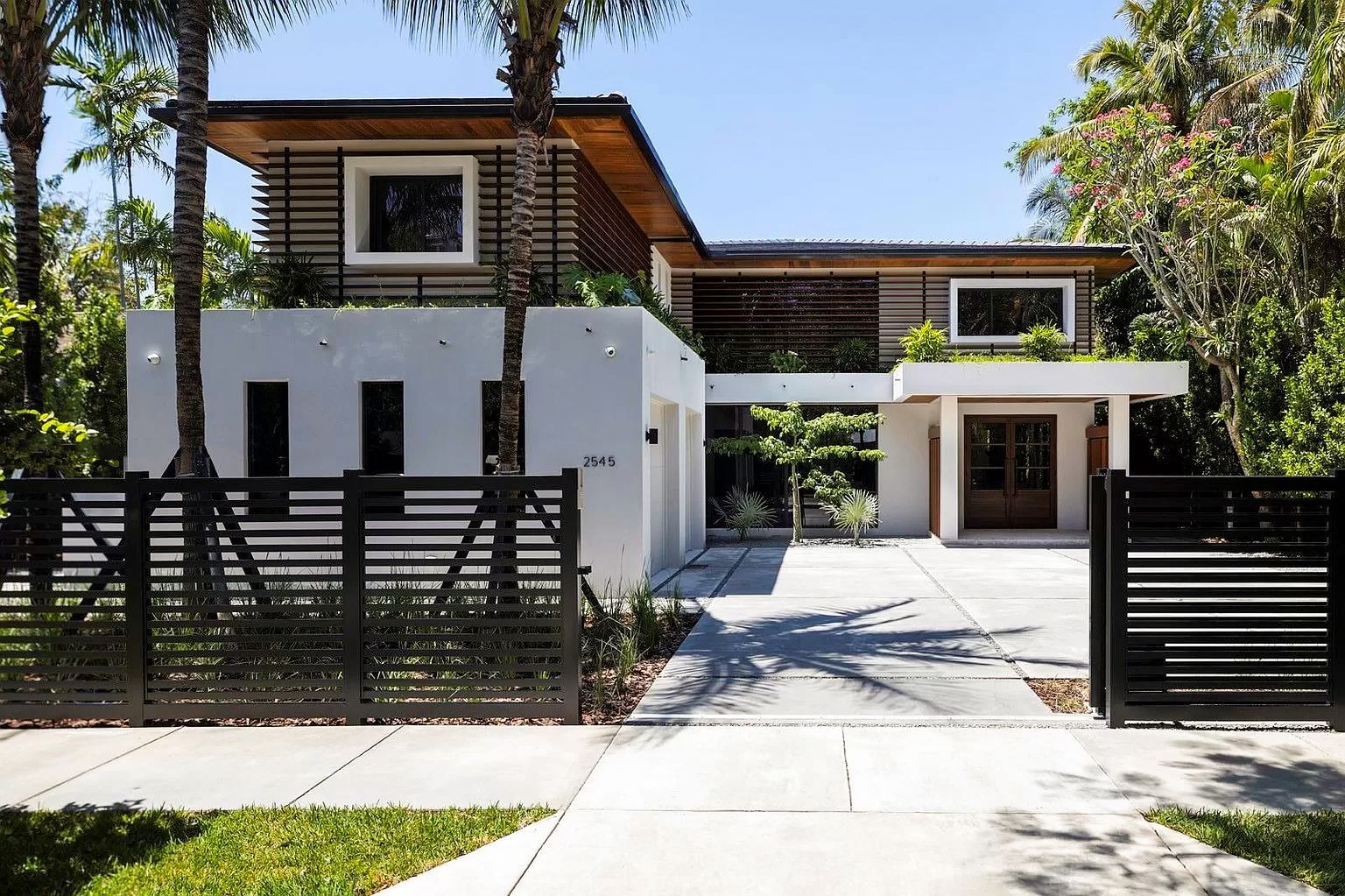 Modern white house with dark horizontal fence, concrete driveway, and lush greenery.