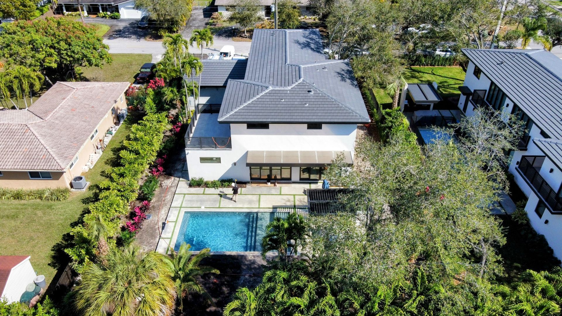 Aerial view of a modern white house with a pool. Lush greenery surrounds the property, sunny day.