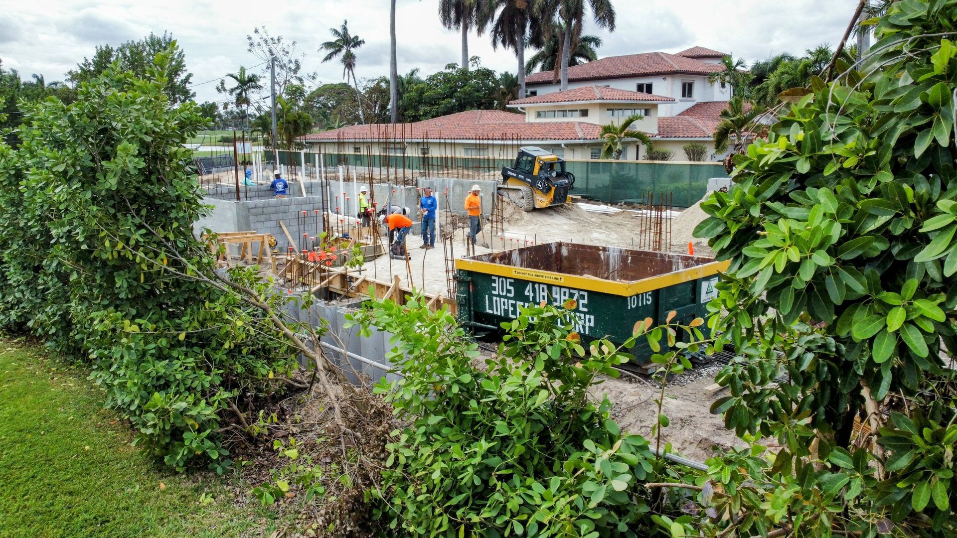 A construction site with a house in the background is surrounded by trees and bushes.