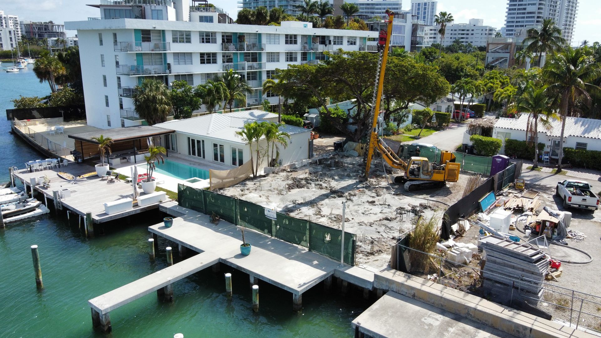Construction site with crane, excavator, and debris, near a dock and waterfront buildings.