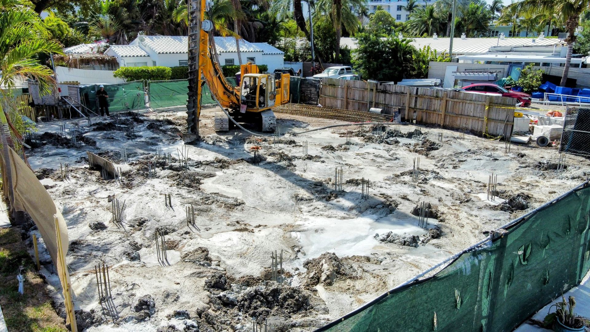 Construction site with excavator. Muddy ground within fenced area; white buildings in background.
