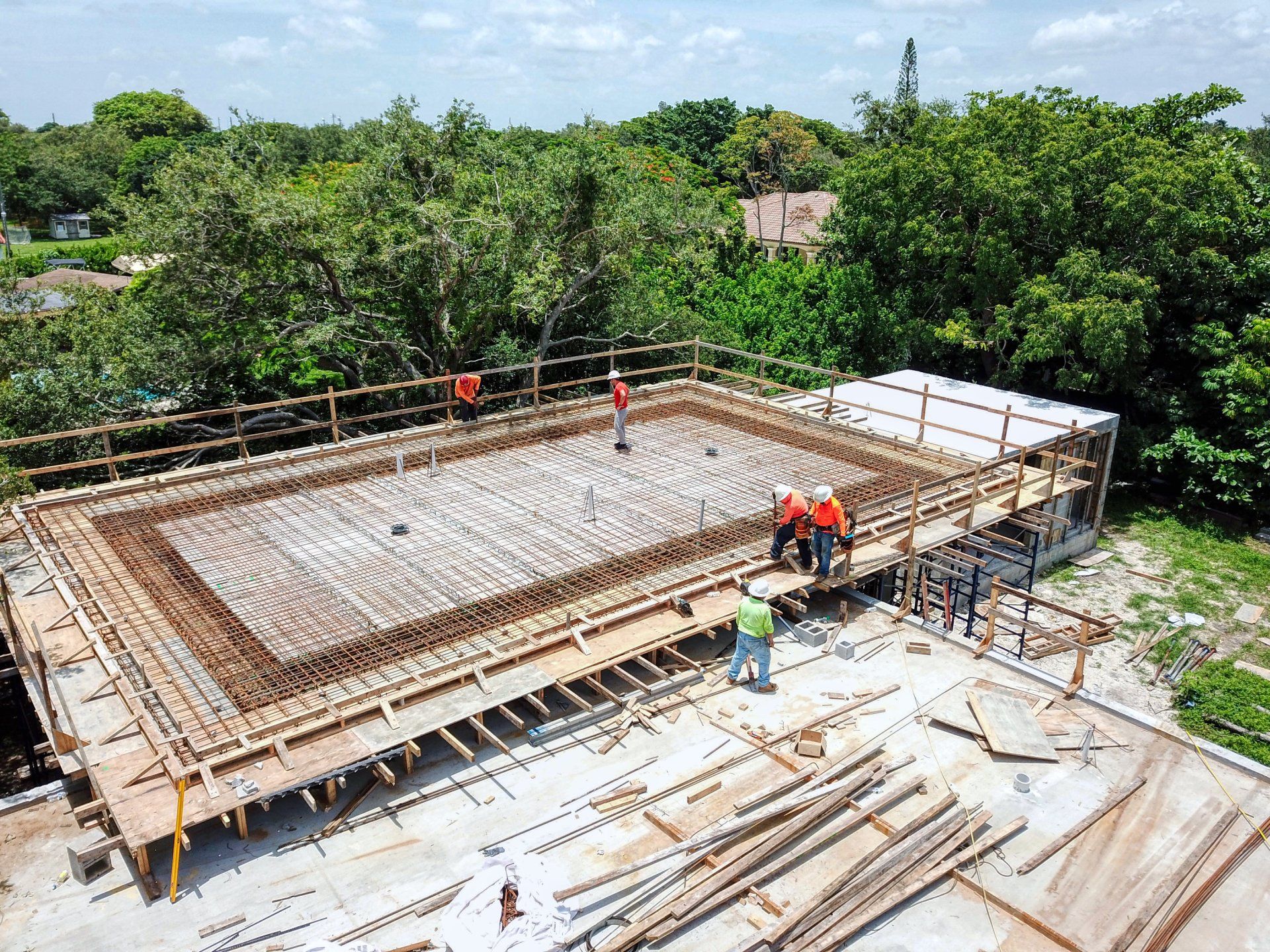 Construction site: Wooden formwork for a rectangular concrete structure. Workers in safety vests are present. Green trees in the background.