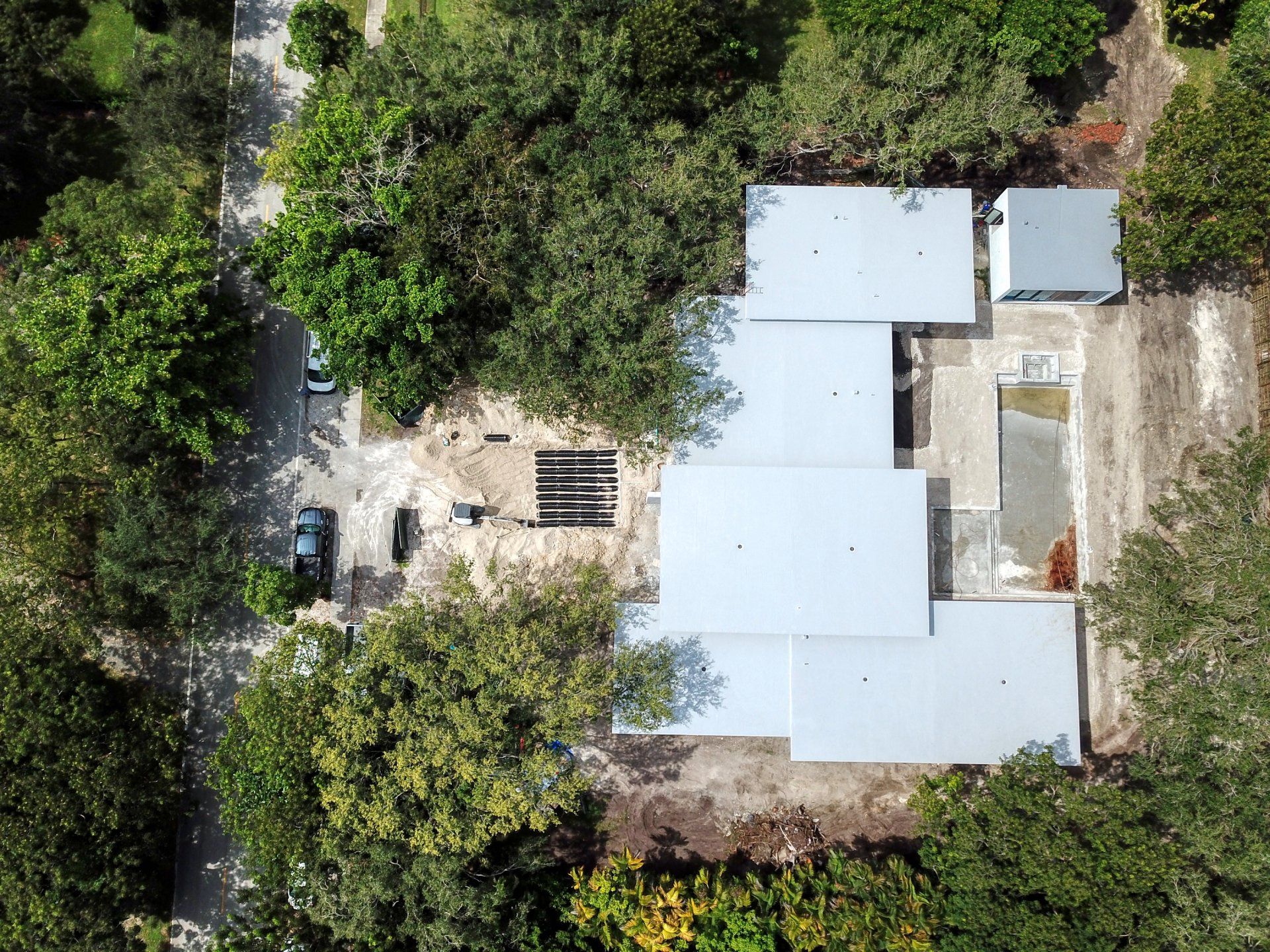Aerial view of a building with a light gray roof surrounded by trees and a road.
