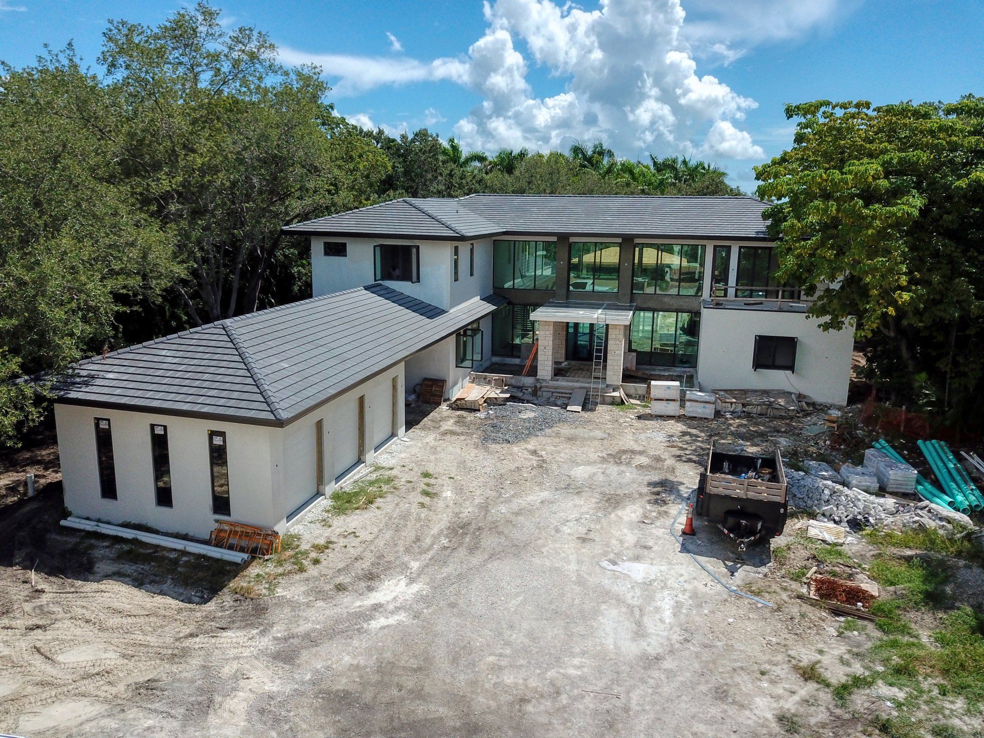 Two-story house under construction with a detached garage. Grey roof, white walls, and a dirt driveway.