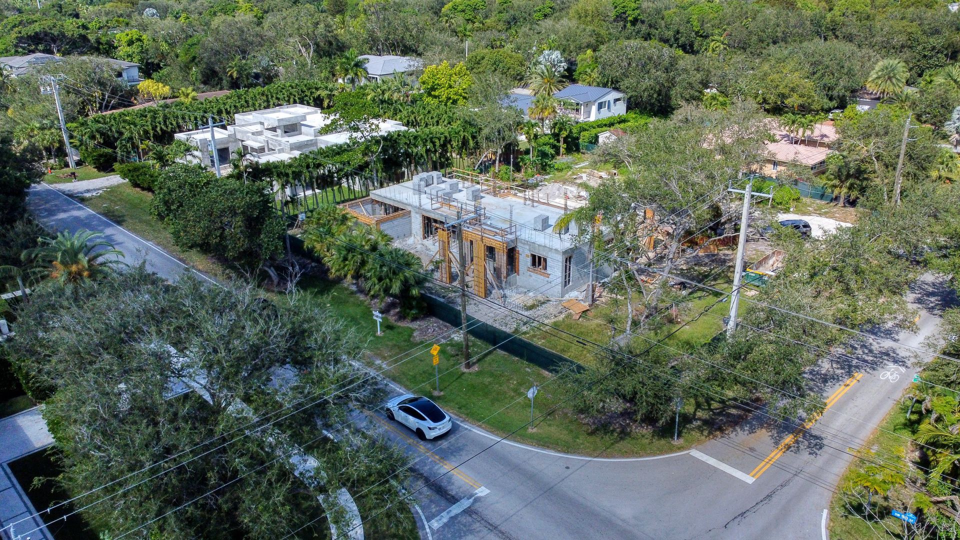 Aerial view of a partially demolished concrete building next to a curved road, surrounded by trees and residential properties.