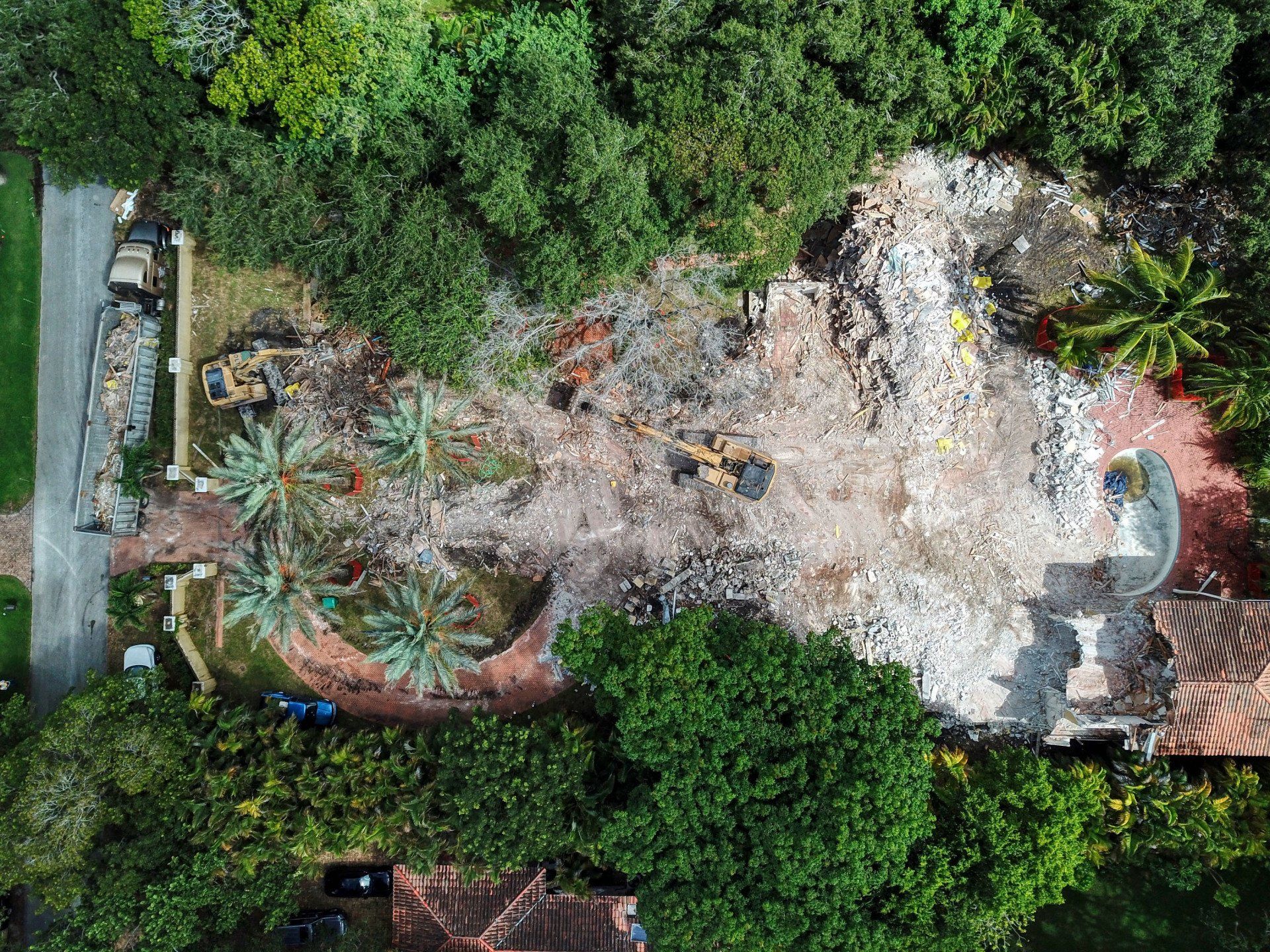 Aerial view of demolished building site, surrounded by trees; excavators and debris visible.