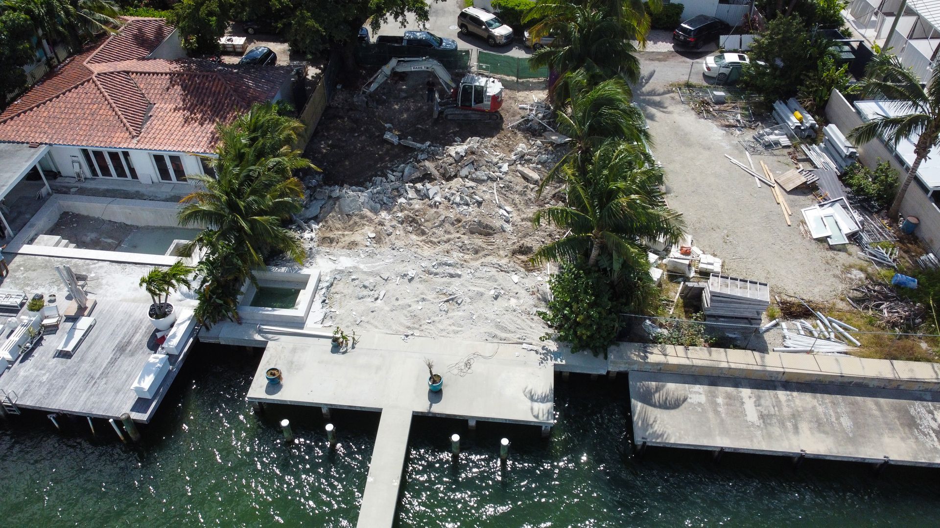 Aerial view of a demolished building site on the water. Debris, docks, and nearby houses are visible.