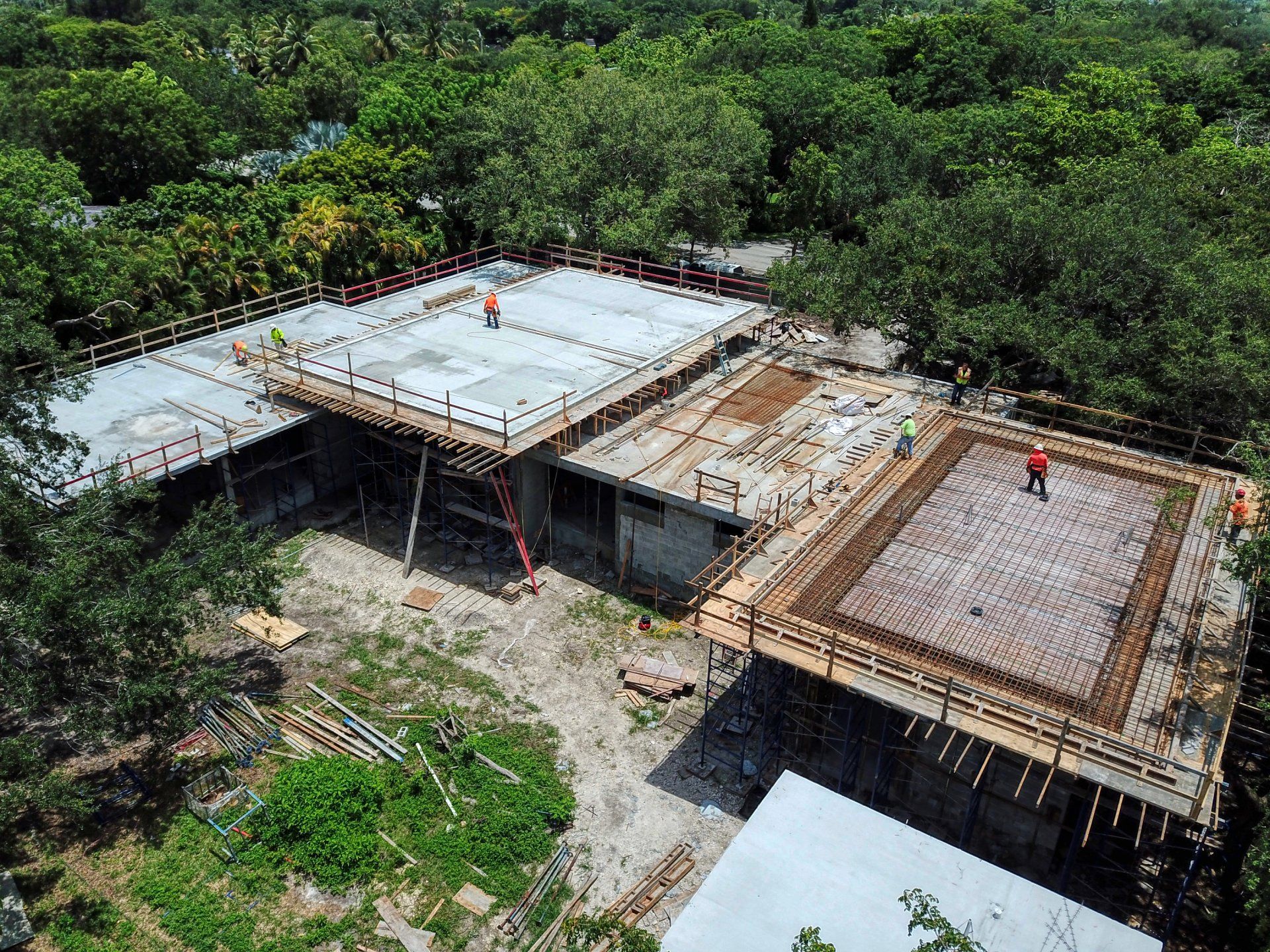 Aerial view of a building under construction, with wooden scaffolding, workers, and concrete forms surrounded by trees.