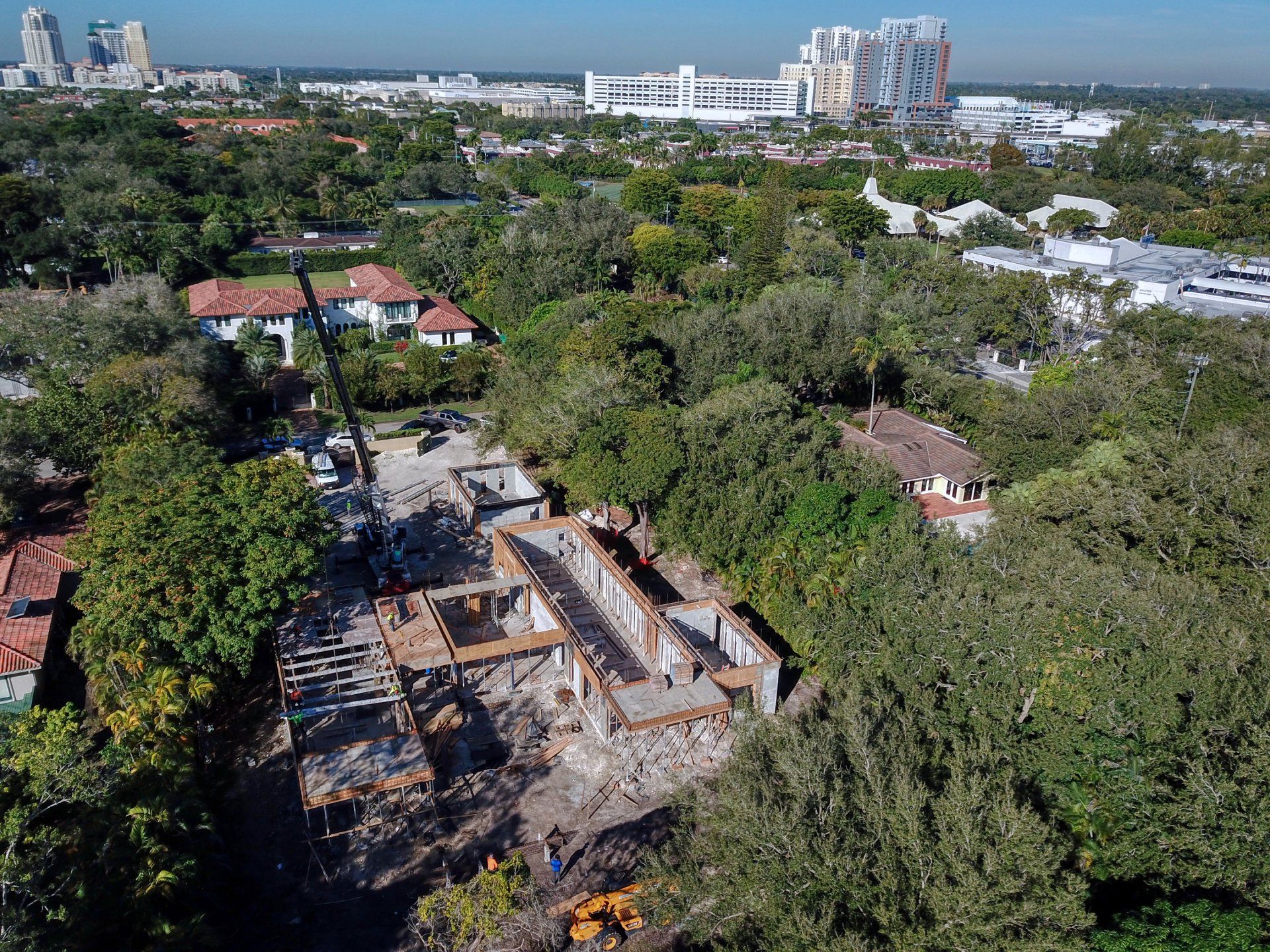 Aerial view of a construction site among trees and houses with a city skyline in the background.