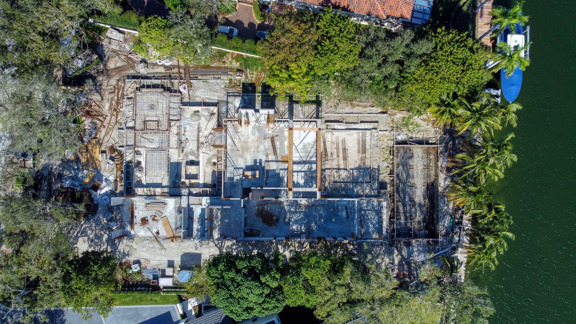 Aerial view of a home demolition site by water. Debris and exposed structure are visible, surrounded by trees.