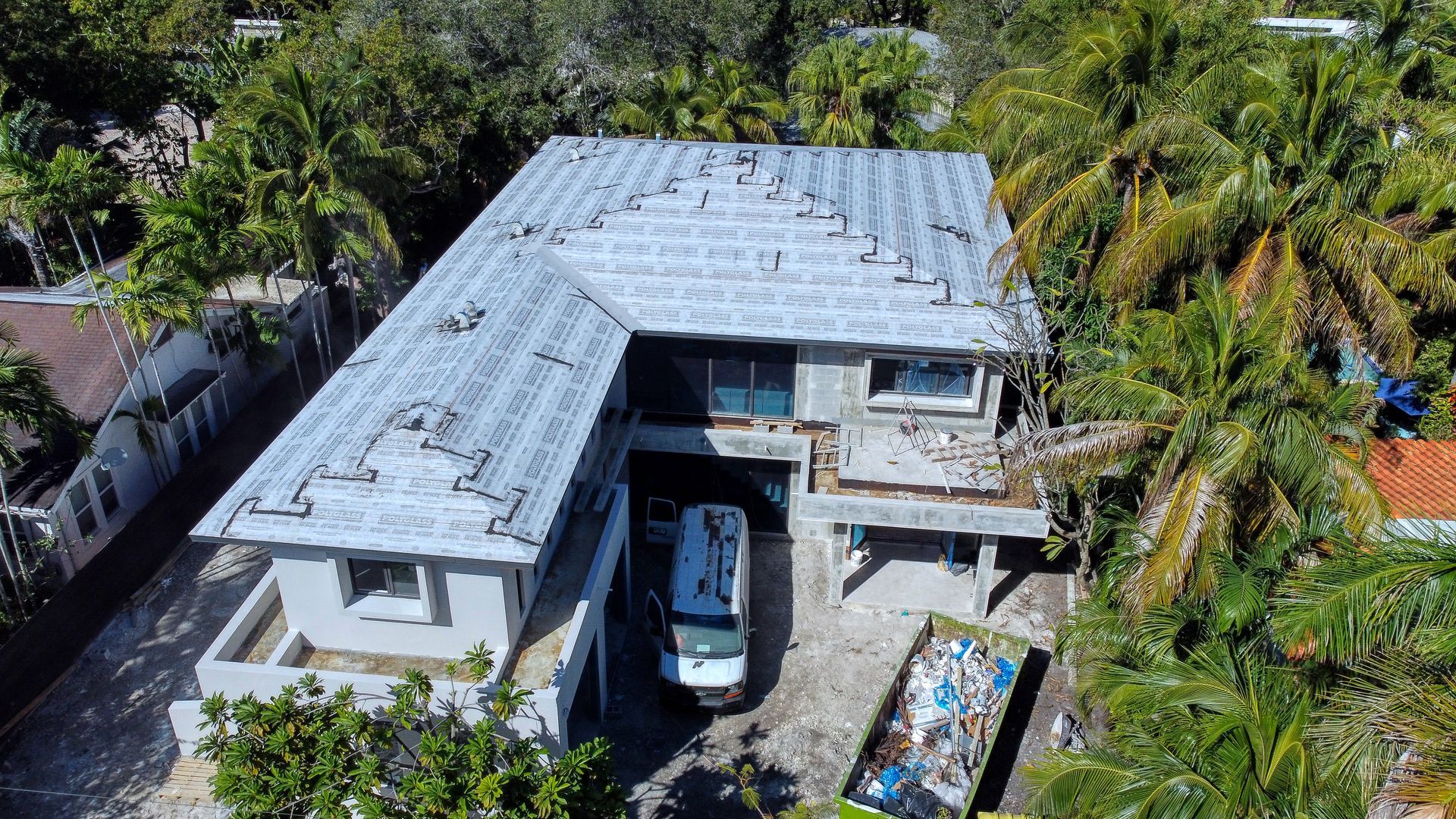 Aerial view of a gray two-story house with roof damage, surrounded by green trees. A vehicle is parked in the driveway.