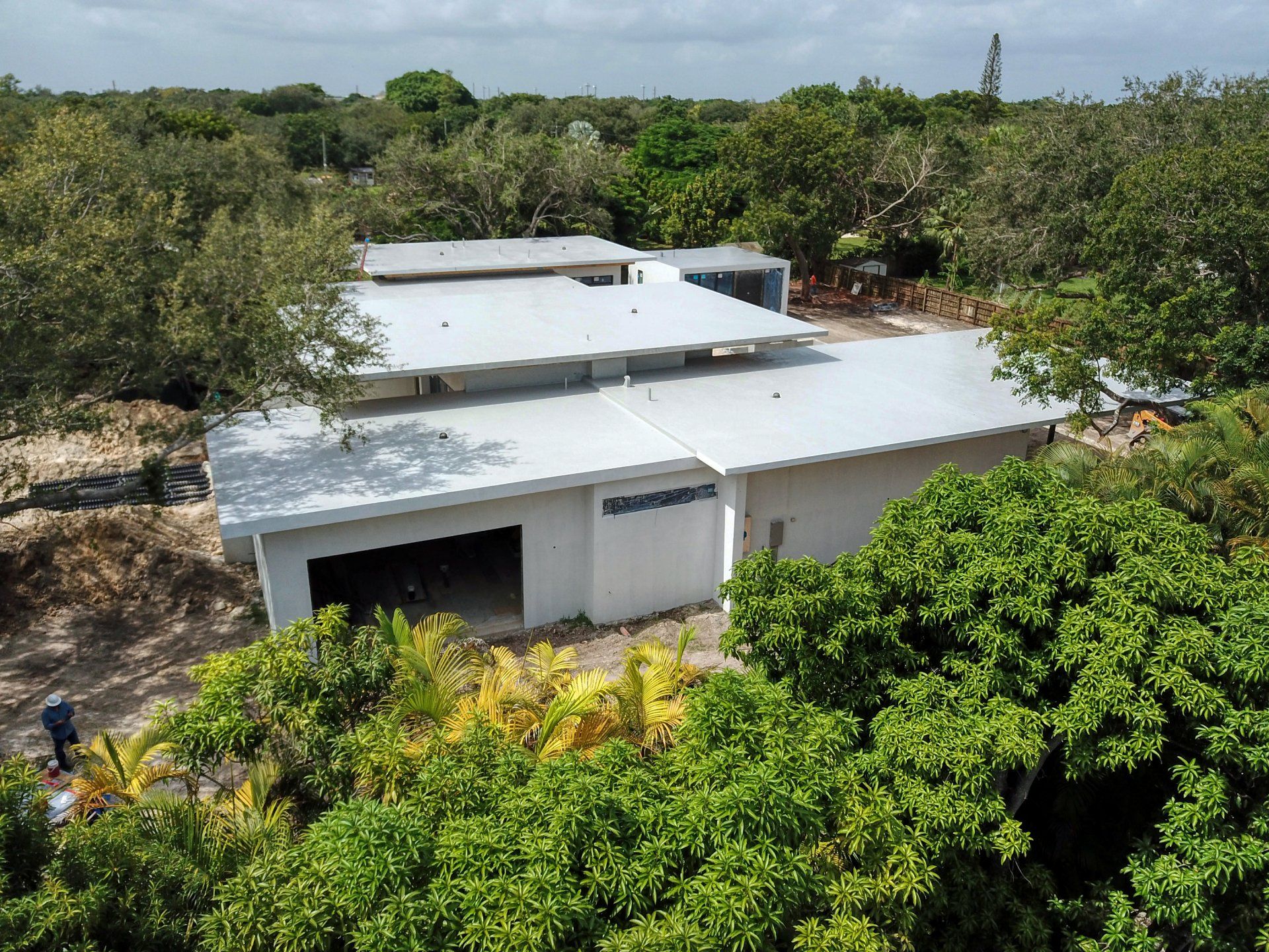 White building with a metal roof surrounded by lush green trees, viewed from above on a sunny day.