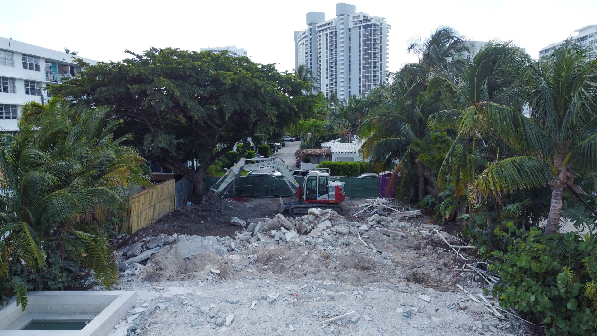 Construction site with excavator amidst rubble, surrounded by trees and buildings.