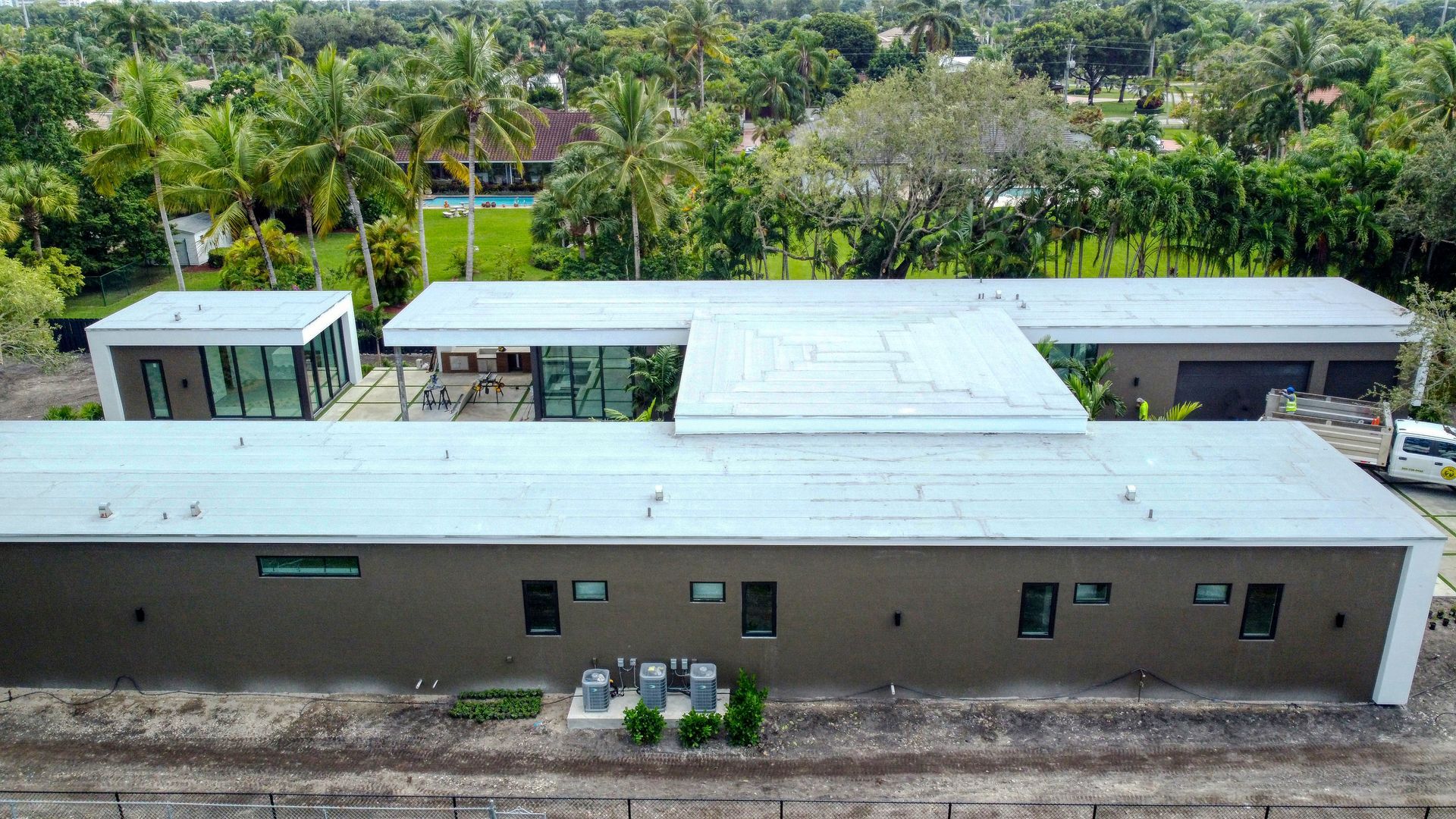 Modern single-story house with flat roof, several sections, and lush green trees in the background.