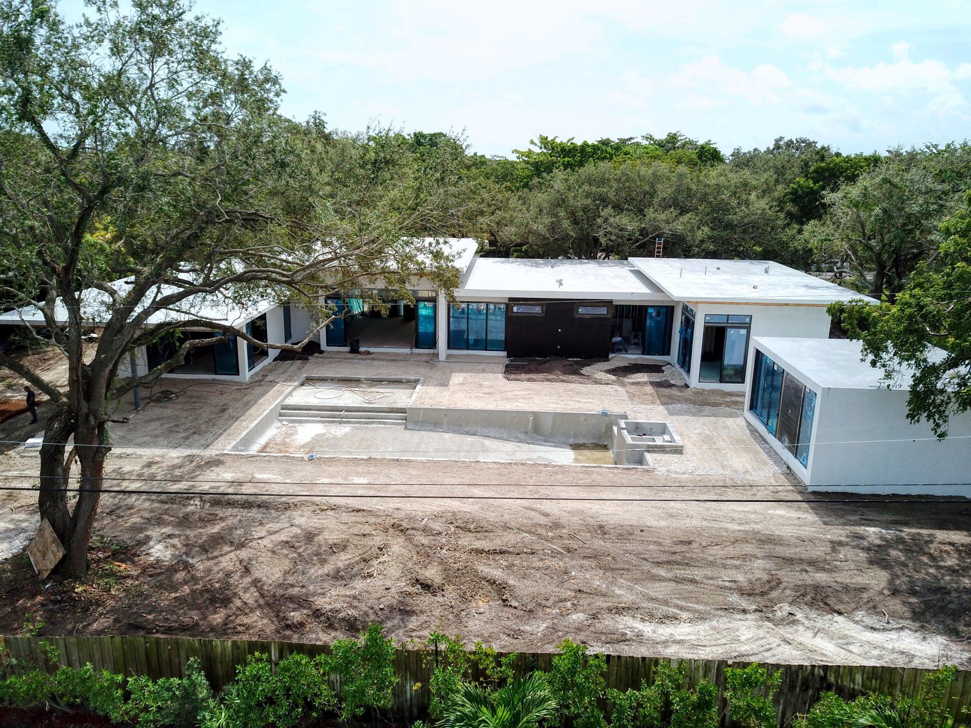 Construction site with a white house, unfinished pool, and surrounding trees.