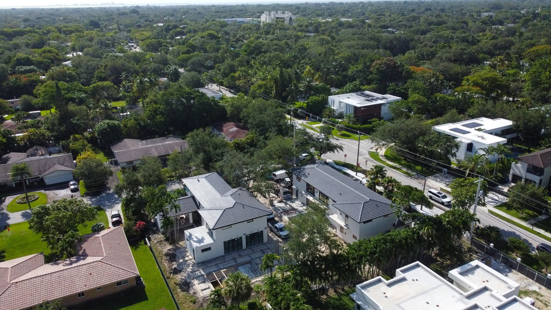 Aerial view of houses with dark gray roofs, surrounded by lush green trees, in a sunny neighborhood.