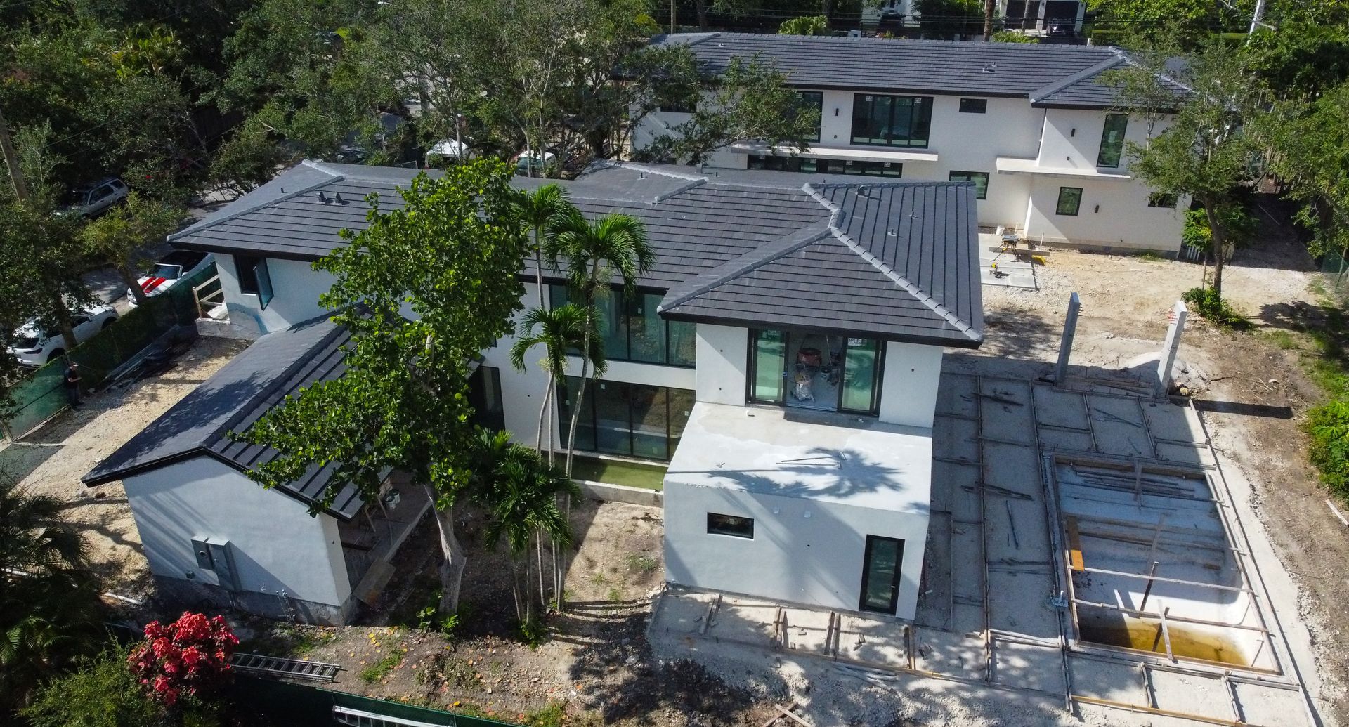 White modern two-story homes with dark gray roofs, surrounded by trees and a dirt lot, under construction.