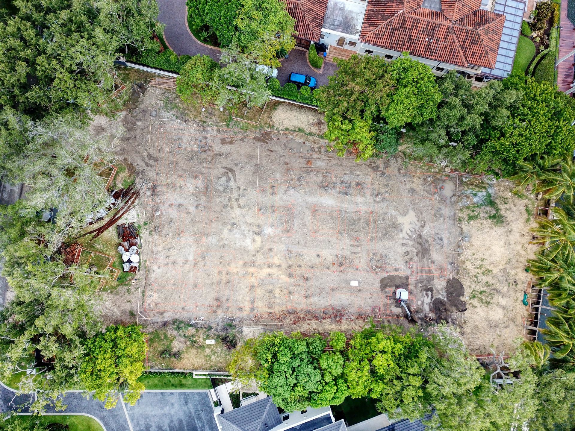 Aerial view of a cleared construction site surrounded by trees and a building with a red tile roof.