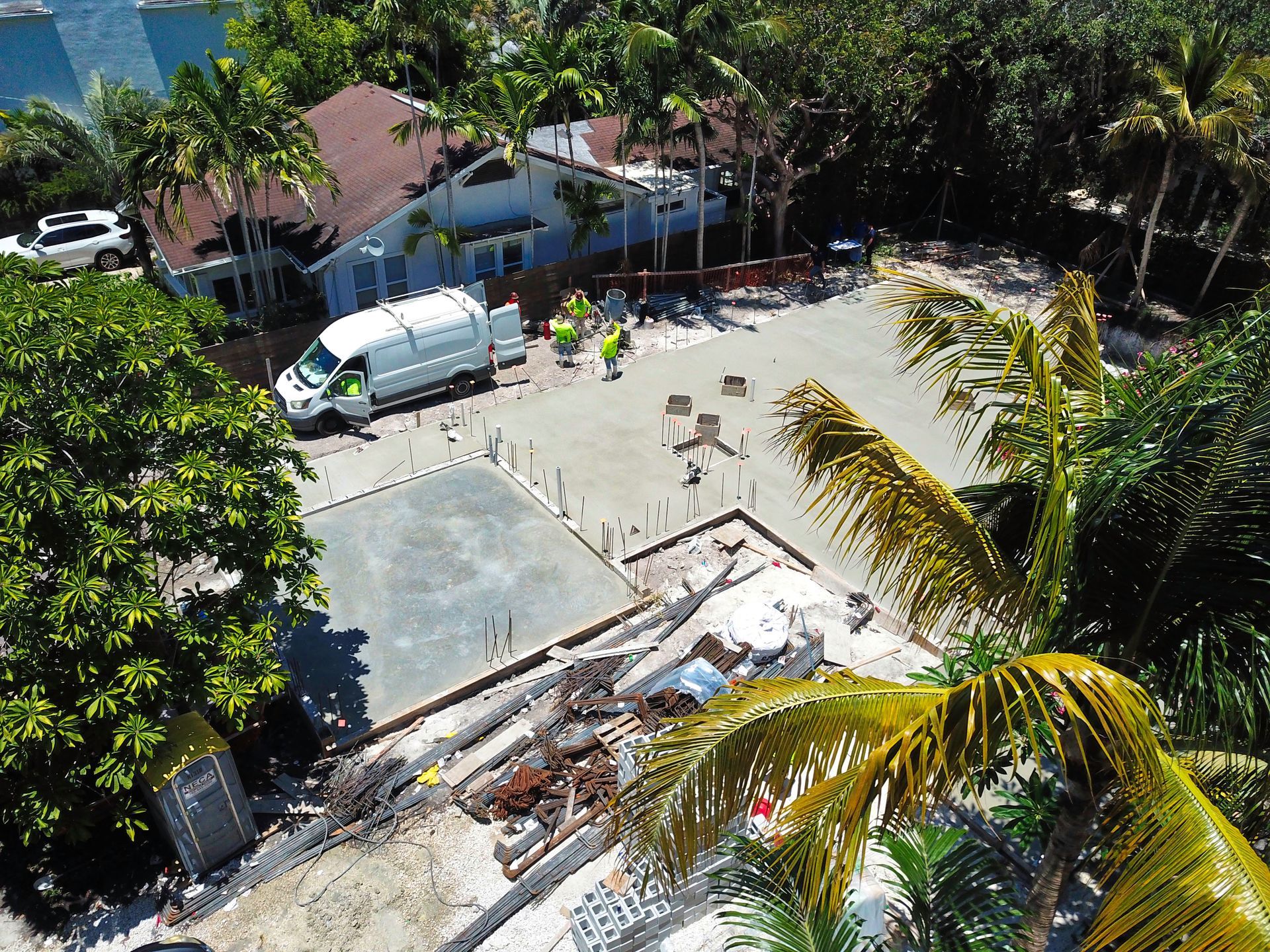 Aerial view of construction site with cement foundation, truck, and building with brown roof surrounded by trees.