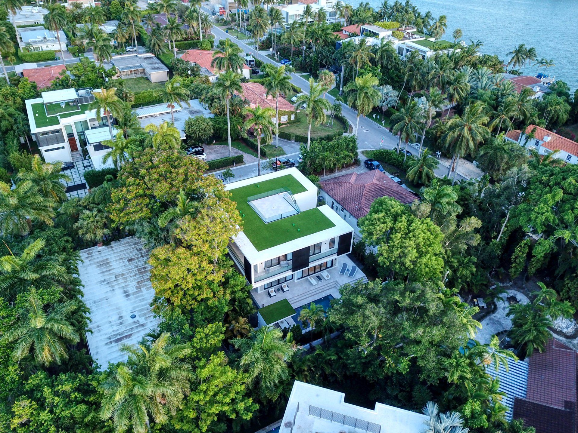 Aerial view of modern white house with a green roof, surrounded by lush green trees and near the water.