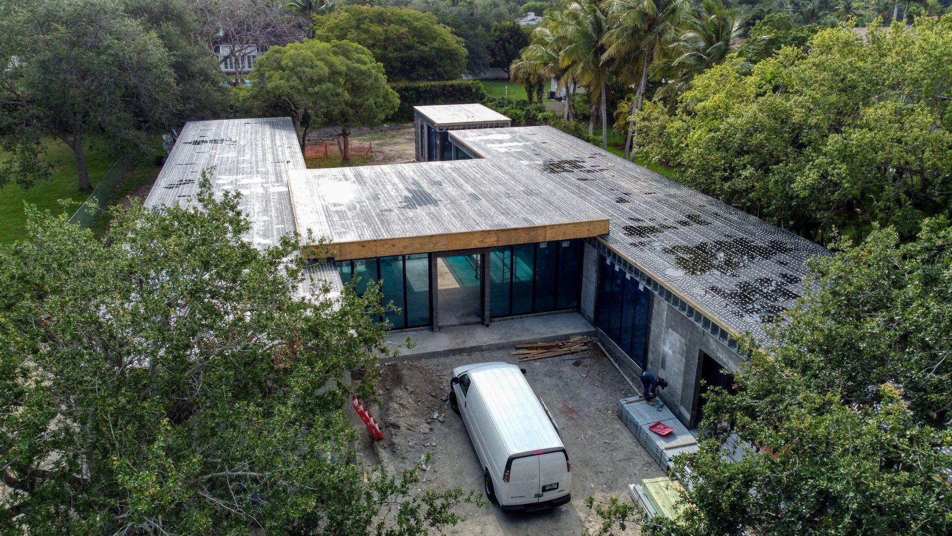 Aerial view of a modern house under construction, surrounded by trees. White van and materials in the driveway.