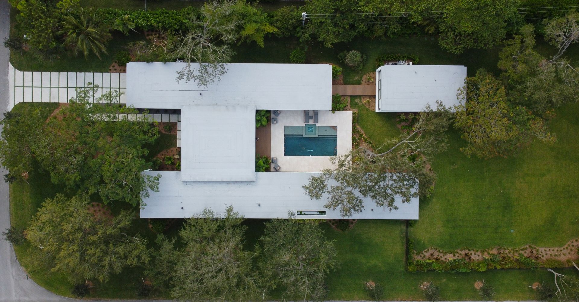 Aerial view of a modern home with a rectangular pool. White roof, concrete driveway, and surrounding green lawn and trees.