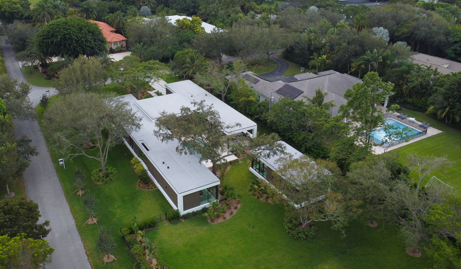 Aerial view of modern white house with a flat roof next to a road and green lawn with surrounding trees.